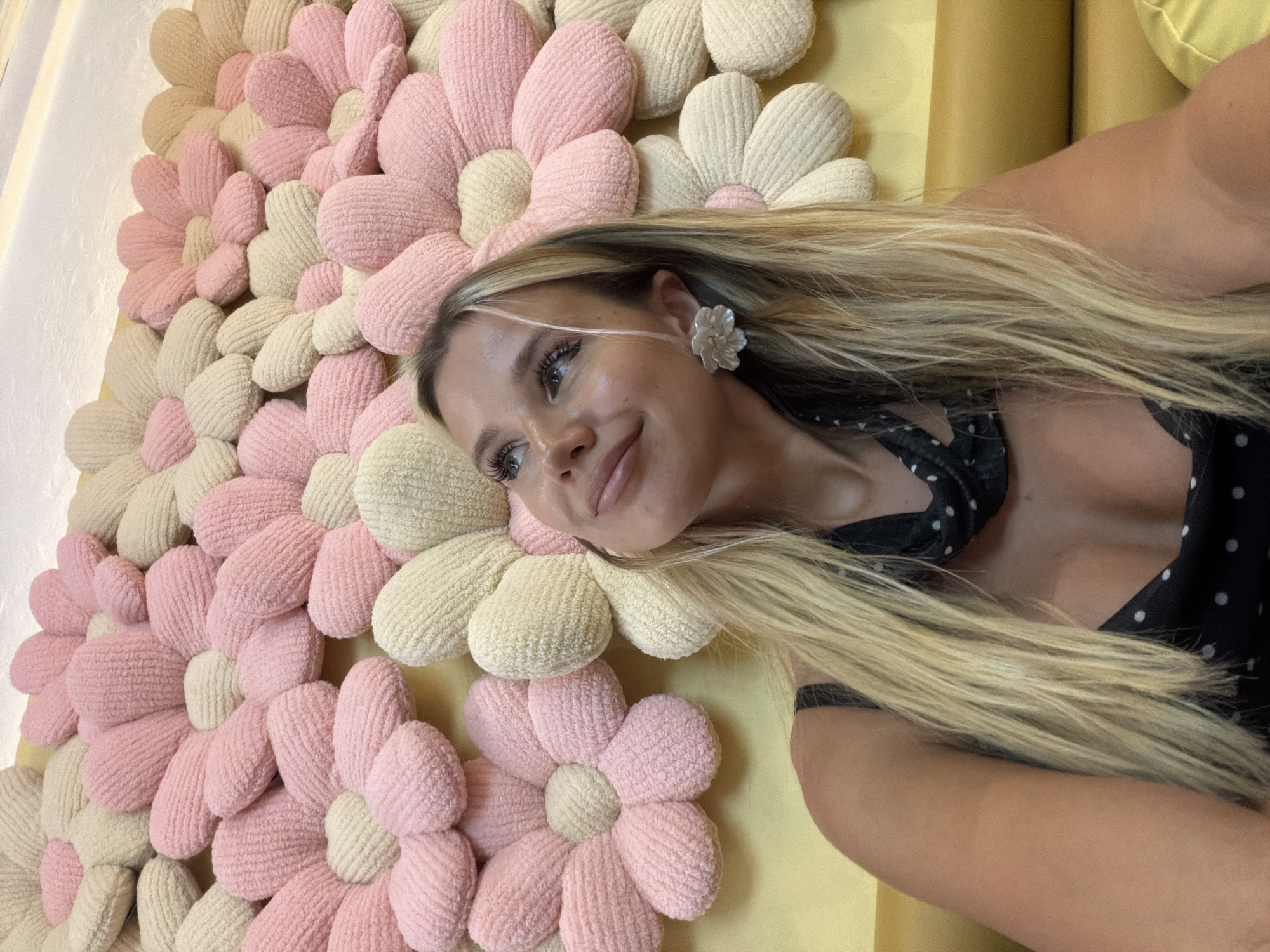 A young woman with blonde hair and large white flower earrings is pictured in front of a wall decorated with large plush flowers in pink and cream colors. She is wearing a black dress with white polka dots and a black and white patterned scarf around her neck. The woman is looking upwards and to her left, with a slight smile on her face. She is seated on a yellow couch. The image appears to be taken indoors, possibly in a restaurant or cafe, with the wall decor suggesting a playful or whimsical setting. The presence of the couch and the woman's attire suggest a casual social setting. There is no indication of the specific location within Phuket City Municipality, Thailand.