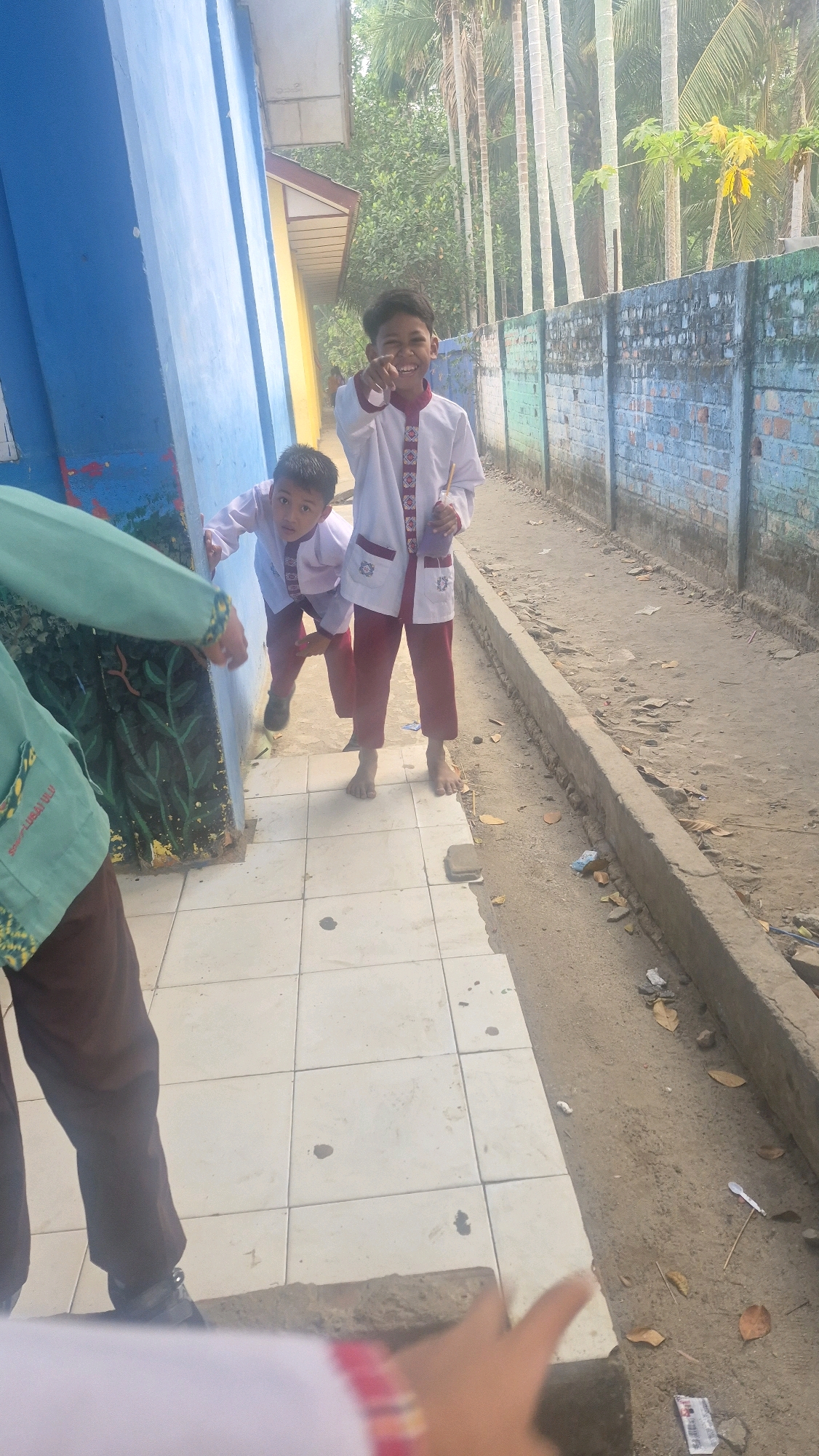 In Putak, Indonesia, two young boys are standing near a blue and yellow building. The boy in the foreground is wearing a white and red uniform, with a red sash. He is pointing at the camera and smiling. The second boy is wearing a similar uniform and is partially obscured by the first boy, but appears to be looking at the camera. A third person, wearing a green uniform, is visible in the lower left corner of the image, with their back to the camera. The boys are standing on a tiled walkway next to a dirt path bordered by a low concrete wall. In the background, there is a brick wall with colorful paint, and tall trees are visible behind it. The environment appears to be outdoors, possibly in a schoolyard or a similar public space.
