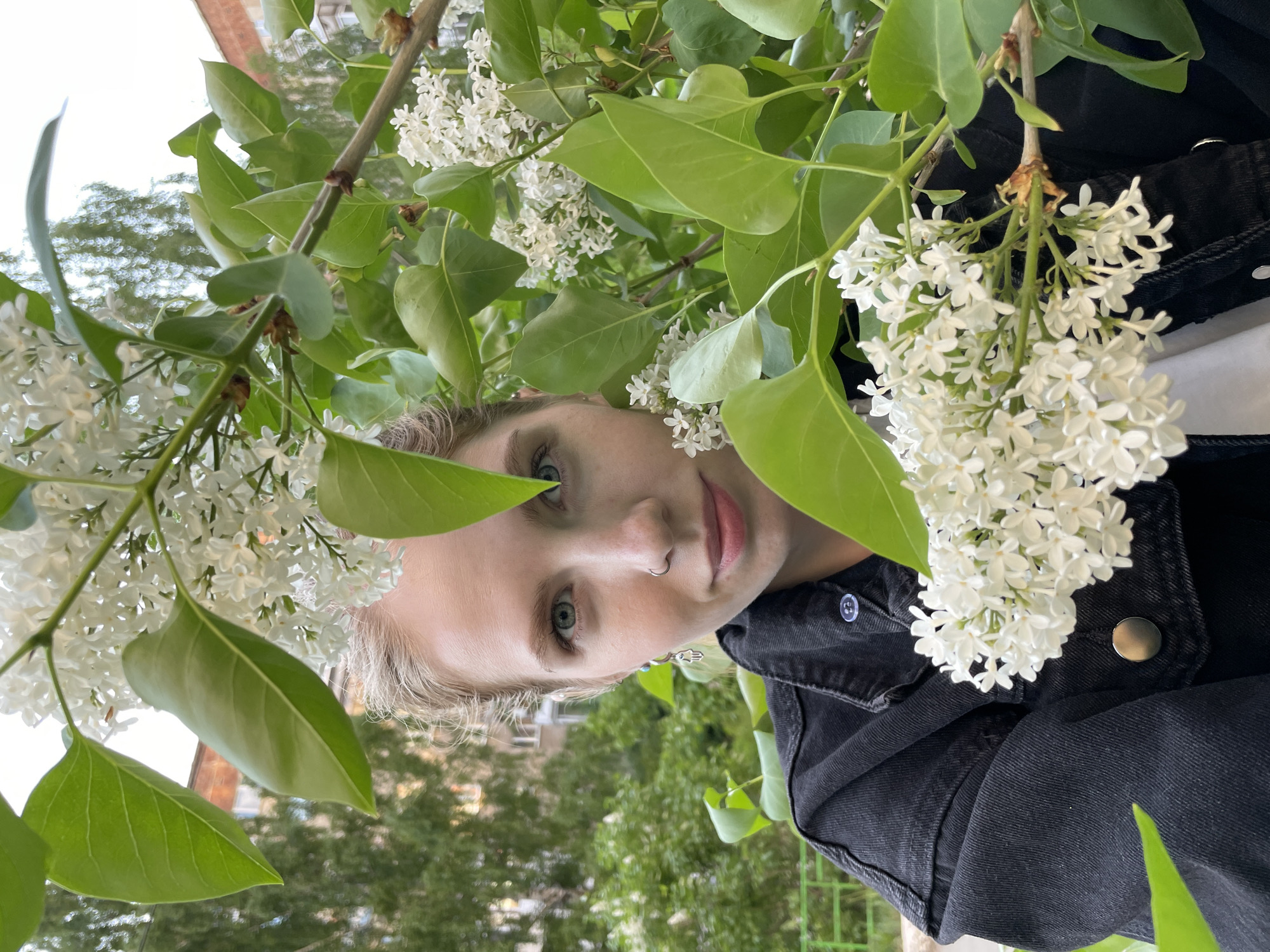A portrait orientation close-up shot shows a person with light blonde hair, fair skin, and blue eyes, partially obscured by clusters of white lilac flowers and green leaves. The person wears a black denim jacket and has a nose ring. The background shows a cityscape with greenery and buildings, potentially within the city of Izhevsk, Russia. The photograph appears to have been taken from a low angle, looking upwards through the flowering branches.