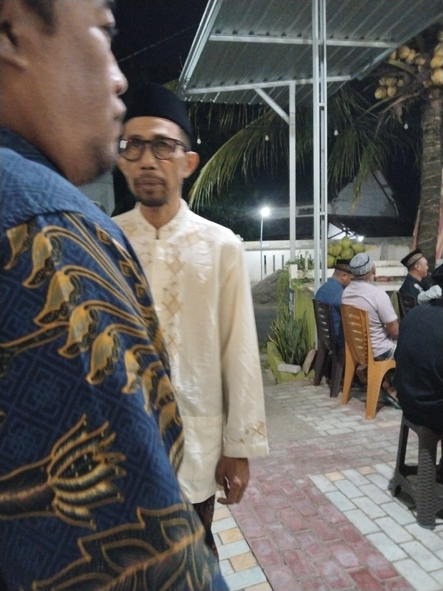 In Prigi, Indonesia, at night, a group of men are gathered outdoors. Two men are in the foreground, the one on the left facing away from the camera and wearing a dark blue garment with a gold floral pattern. The man to his right, facing the camera, wears a white, long-sleeved shirt and a black cap, and glasses. In the background, several men are seated on chairs, one in a white shirt and a grey cap, another in a blue shirt, and others are partially visible. A coconut tree with a cluster of coconuts is visible to the right, and a structure with a metal roof is overhead. The ground is paved with brick tiles.