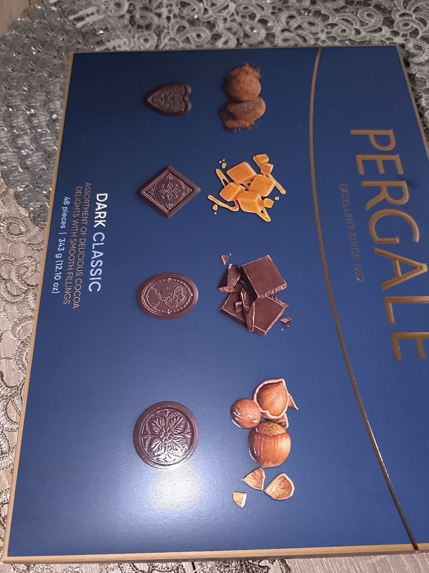 A dark blue chocolate box, branded "PERGALE" in gold capital letters vertically on the right side, lies on a light-colored, patterned, lacy textile surface. The box features gold trim along its edges. Horizontally aligned on the left side of the box, in gold lettering, are the words "DARK CLASSIC" followed by "ASSORTMENT OF DELICIOUS COCOA DELIGHTS WITH SMOOTH FILLINGS." Below this text, also in gold, it reads "48 pieces | 343 g (12.10 oz)." The phrase "EXCELLENT SINCE 1952" is inscribed vertically below the "PERGALE" logo.
The box cover is adorned with illustrated depictions of various chocolate types and ingredients:
* In the upper left, a heart-shaped dark chocolate piece with an embossed design.
* Below it, a square dark chocolate piece with an embossed snowflake-like pattern.
* Further down, a circular dark chocolate piece featuring an embossed profile, possibly a woman's head.
* At the bottom left, a circular dark chocolate piece with an intricate floral or mandala embossed design.
* Near the top right-center, two irregular, spherical chocolate truffles or pralines, appearing to have a powdered coating.
* Below the truffles, several small, irregular, golden-orange cubes, resembling caramel, with depicted drizzles.
* Further down, a small pile of broken pieces of dark chocolate.
* At the bottom right-center, a cluster of hazelnuts, some shelled, some unshelled, accompanied by broken shell fragments.
The patterned textile surface beneath the box displays a repeating circular or floral motif. The overall scene is illuminated by indoor lighting, creating a subtle sheen on the box's surface. The geographical location in Dercekliai, Lithuania, cannot be determined from the visual information provided in the image.