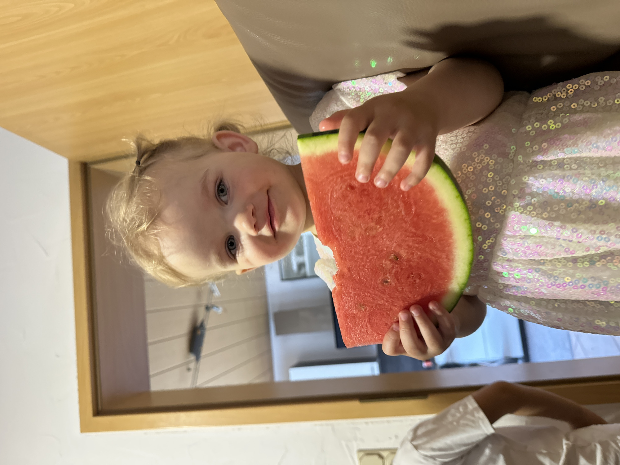 A young girl with blonde hair is holding and eating a slice of watermelon. She is wearing a white dress with sequins. The background shows an interior with a wooden door frame and a glimpse of another room. It is unclear if this image was taken in Niederquembach, Germany.
