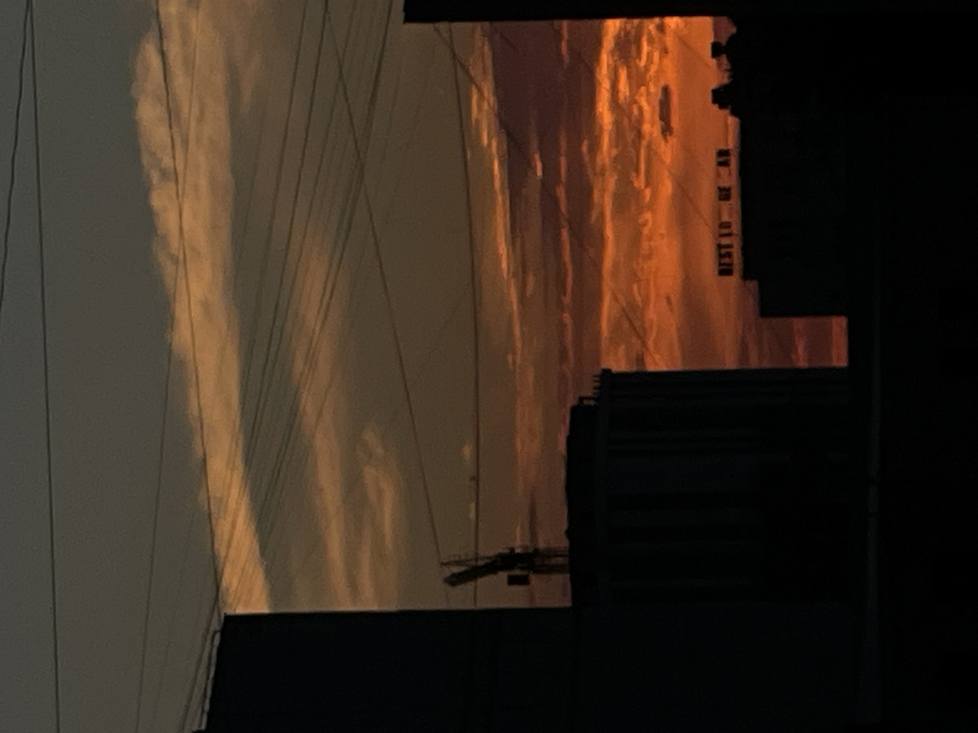 The image shows a cityscape at sunset, with the sky ablaze in oranges and reds. Silhouetted buildings and a crane dominate the foreground, with power lines crisscrossing the frame. A sign on top of one of the buildings reads "BEST LO UN AR," which is likely an advertisement. The overall impression is one of urban life against a dramatic natural backdrop. The location is identified as Aktau, Kazakhstan.