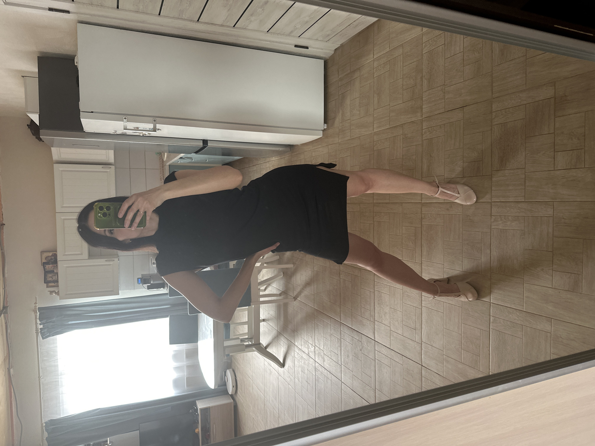 An indoor mirror selfie captures a woman posing in what appears to be a kitchen or dining area. The image is rotated 90 degrees clockwise, showing the scene on its side.
The woman, with dark hair, is wearing a black dress and light-colored, possibly ballet-style, flats. She holds a green-cased smartphone to take the picture, partially obscuring her face. A ring is visible on her left hand. She is positioned with one leg extended slightly back, hands on her hips, looking towards the phone.
The background, reflected in the mirror, reveals elements of a home. To her right (upper left in the rotated image) is a white refrigerator and white upper kitchen cabinets, with a metallic range hood visible beneath them. To her left (lower right in the rotated image), a dining table with white legs and chairs is visible, along with a circular robotic vacuum cleaner on the tiled floor nearby. A large window with grey curtains lets in ample light, suggesting daytime. The flooring throughout the visible area is light brown tile with a rectangular pattern. The ceiling appears light-colored with a subtle texture. No discernable text is visible.