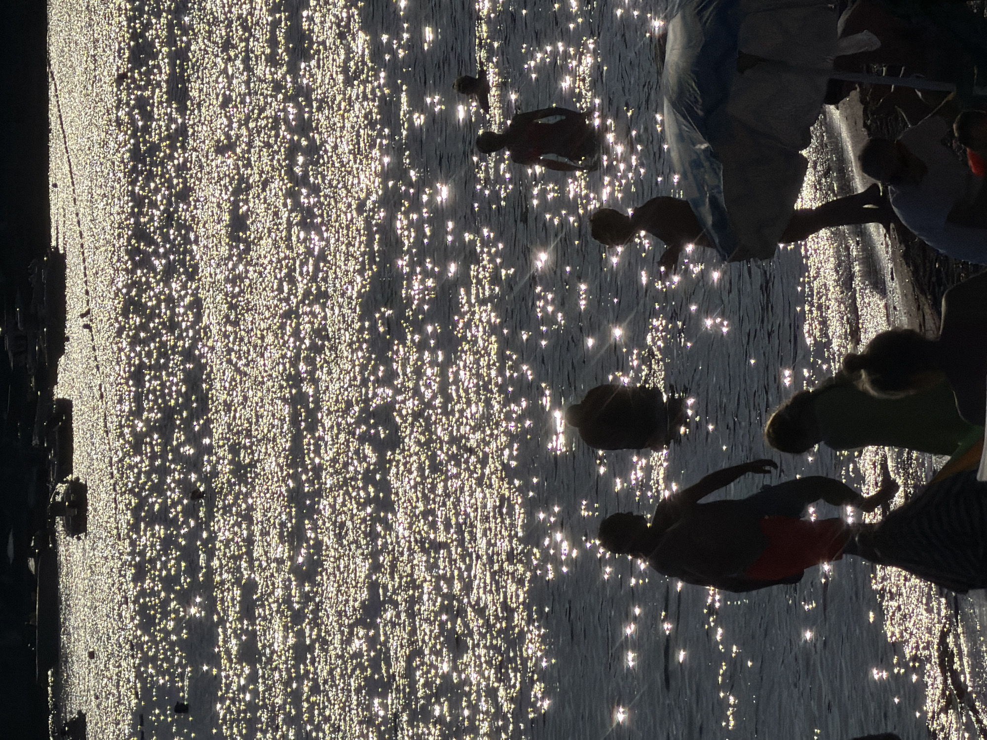 The image shows a sun-drenched scene at the Petrovac centar, Montenegro beach. The water sparkles with a multitude of reflections from the sun. In the water, several people are visible: one person is standing in the water, another is further out, and two more are in the foreground partially submerged. On the shore, some individuals are relaxing under a large beach umbrella. Boats are moored in the distance, silhouetted against the bright water. The overall impression is one of a lively and sunny day at the beach.