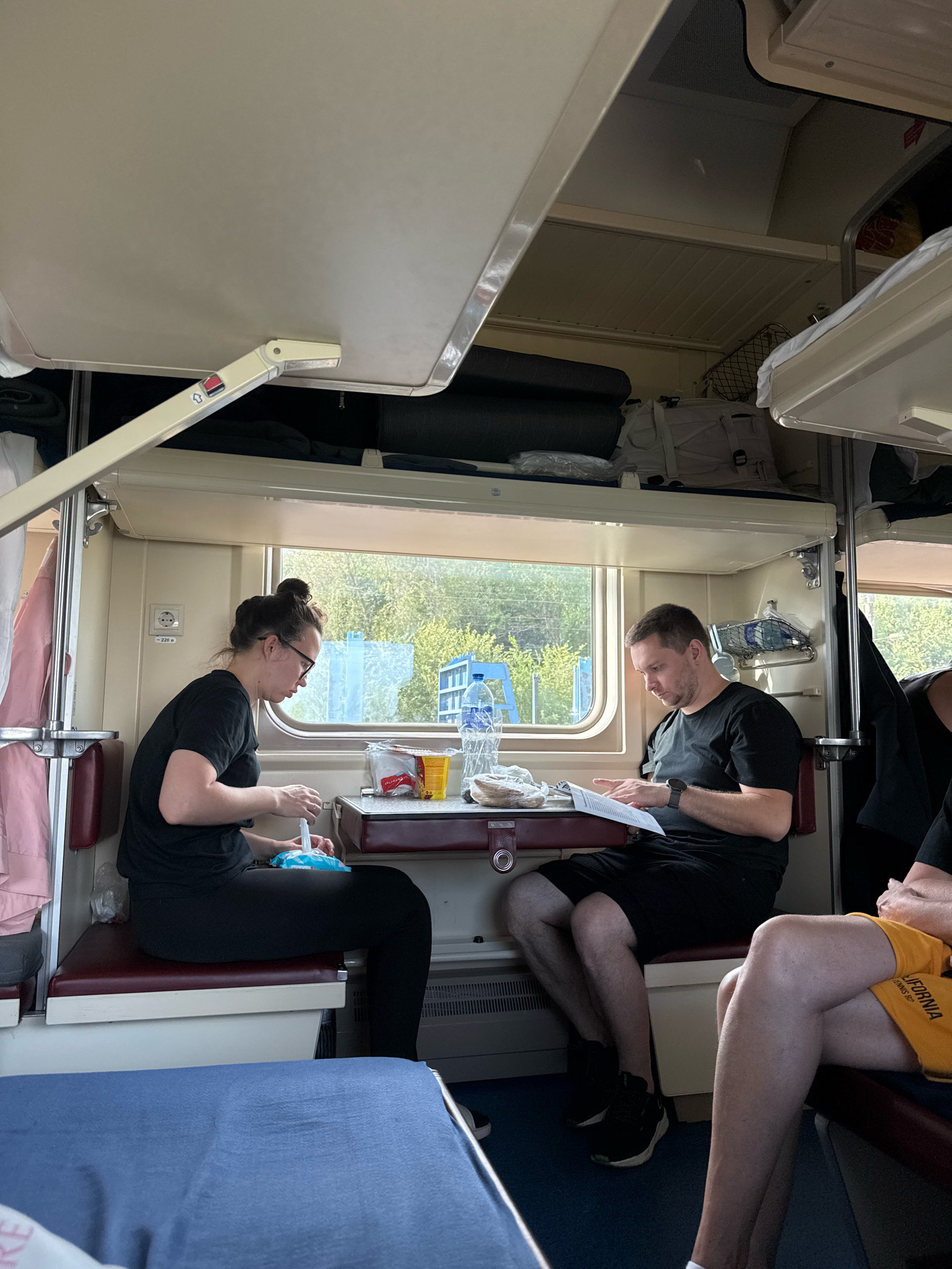 This image depicts the interior of a train compartment, likely during daylight hours. Two individuals, a woman and a man, are seated across a small table from each other. The woman, on the left, is wearing glasses and a black t-shirt with black leggings. She appears to be holding a white object, possibly a tissue or wipe, and is focused on something in her lap. The man, on the right, is wearing a black t-shirt and black shorts. He is looking down at some papers on the table, with his left hand holding them open and his right hand resting on them.
The compartment has a window on the right side, through which trees and some buildings can be seen, suggesting a landscape outside. Inside, there are overhead storage compartments and bunk beds. A blue blanket is visible in the foreground, and a bottle of water and some food items are on the table. The overall environment is utilitarian and typical of train travel. There is no indication that this scene is in Rostov-on-Don, Russia.