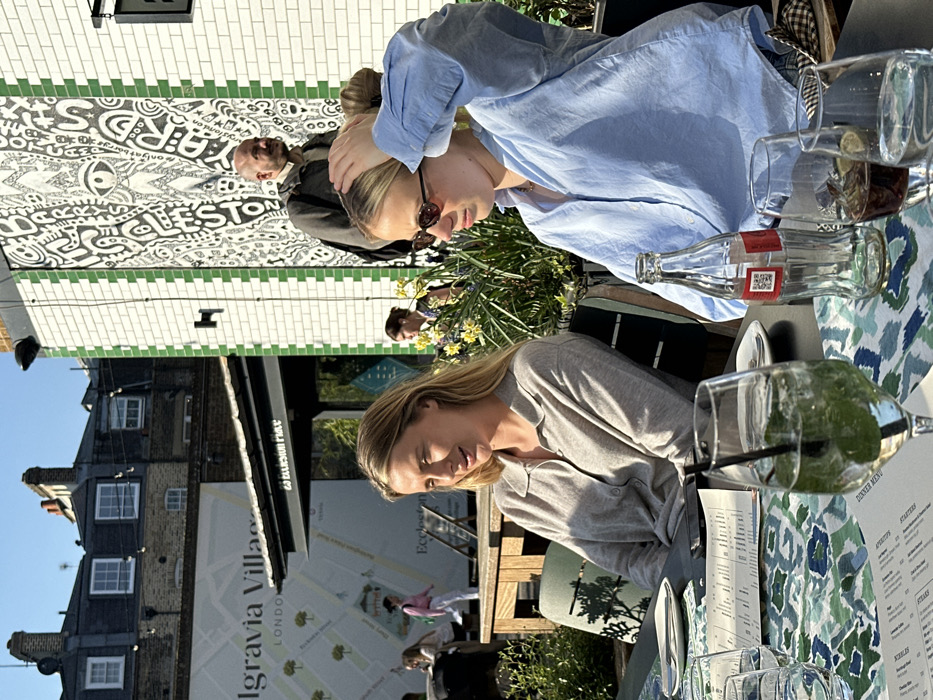 The image displays an outdoor dining area in Belgravia Village, London, United Kingdom, specifically identifiable by a banner stating "Belgravia Village London" and a sign for "25 Eccleston Pl". In the foreground, two blonde women are seated at a dark table with a blue and green patterned runner. The woman on the left wears a light grey collared shirt and is looking left with a slight smile. A tall glass with a green drink, mint leaves, and a dark straw is positioned in front of her. The woman on the right, wearing a light blue button-up shirt and sunglasses, has her hands raised towards her hair and is looking downwards. An empty glass Coca-Cola bottle with a red label and QR code, two additional glasses containing dark liquids and ice (one with a lime wedge), and several menus are on the table.
Behind the seated women, a bald man with a beard, wearing a dark jacket, stands facing left, positioned in front of a white tiled wall featuring a black and white abstract mural with green vertical border tiles, containing text including "ECCLESTON YARD". Further in the background, beyond an awning, are additional blurred figures and older brick buildings with multiple windows and chimneys under a bright, clear sky. Green foliage and yellow flowers are visible in planters behind the table. The scene is illuminated by strong direct sunlight, creating distinct shadows.