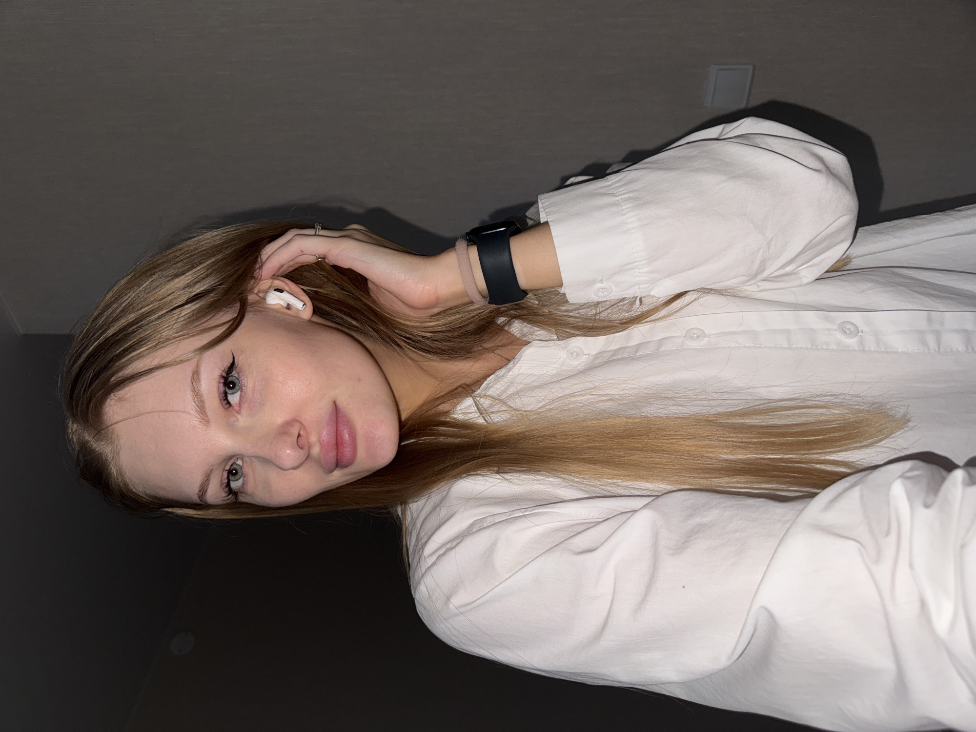 A young Caucasian woman with long blonde hair and light blue eyes is shown in a medium close-up shot. She is wearing a white button-down shirt and has a white wireless earbud in her right ear, with her hand resting on it. She is also wearing a black smartwatch with a light pink band on her left wrist and a ring on her left ring finger. The background is a plain gray wall, dimly lit, with a white electrical outlet visible to the right. The setting appears to be indoors, likely a residential or hotel room, and there is no indication of the city of Perm, Russia.