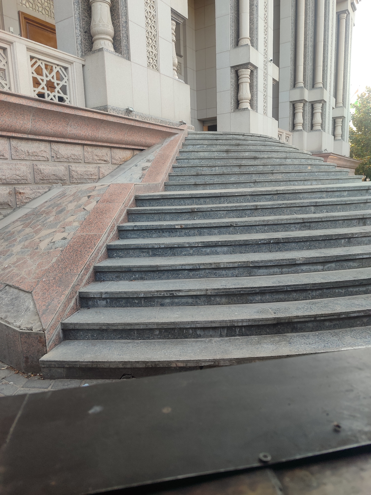 The image shows a wide, curved staircase leading up to a building in Dushanbe, Tajikistan. The staircase is made of gray stone slabs, and there is a ramp on the left side. The building appears to be made of light-colored stone and has ornate columns. The surrounding area is paved with stone tiles, and there are some trees and foliage visible on the right side. A dark, metallic object is in the foreground, partially obscuring the bottom of the staircase. The lighting suggests it is daytime. No people are clearly visible in the scene.