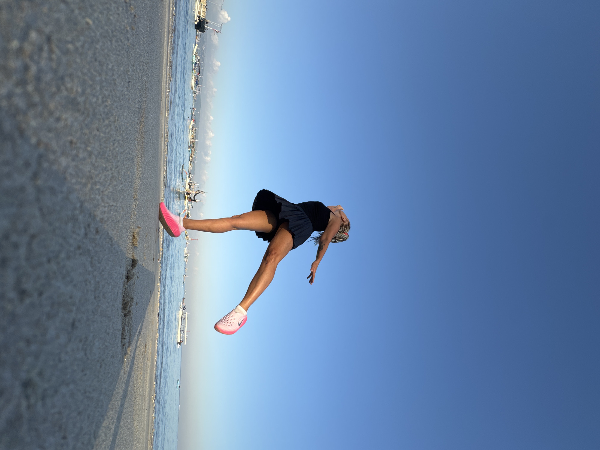 An athletic woman with blonde hair in a black tank top and dark blue skirt is captured mid-air, appearing to leap or dance on a sandy beach. She wears bright pink athletic shoes. Her arms are outstretched, and her head is tilted back. The background features a calm, blue ocean with numerous boats of various sizes scattered across the water. The sky is a clear, deep blue, indicating a bright day. The foreground shows the sandy texture of the beach with a shadow cast from the woman. The scene is located on a beach in Kuta, Indonesia, suggested by the style of the boats and the general coastal environment.