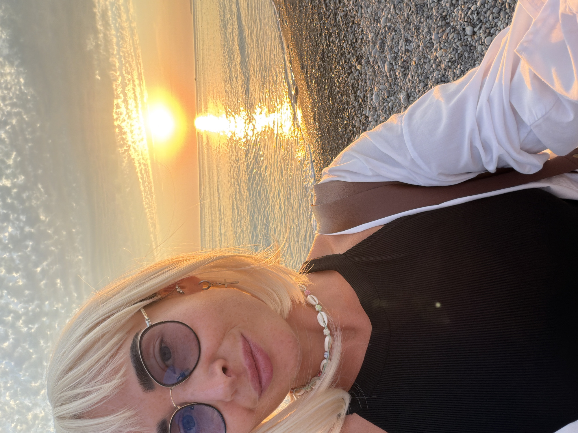 This is a vertical, close-up selfie of a blonde woman with bangs, wearing round sunglasses with blue lenses, a black ribbed tank top, a necklace made of small shells, and a white button-up shirt. She is on a pebble beach with the Black Sea and a sunset in the background. The sunset casts a golden glow over the water and sky, creating a reflection on the sea. The location appears to be Sochi, Russia, given the presence of a pebble beach and the distinct sunset over the Black Sea.