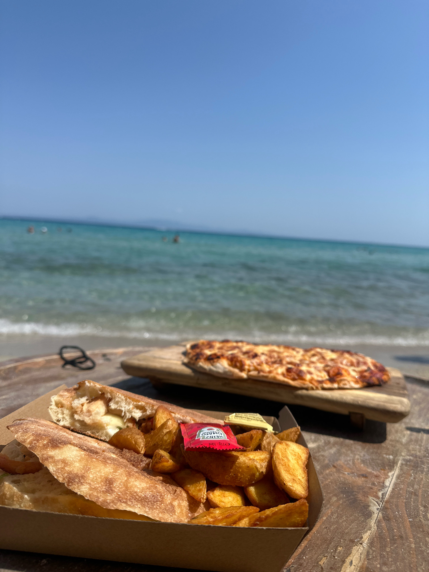The image depicts a meal served on a rustic wooden surface at a beach in the Kassandra Municipal Unit, Greece. In the foreground, a cardboard container holds a sandwich cut in half, revealing filling and a side of potato wedges. Two small condiment packets, one red for ketchup and one yellow for mustard, are placed on top of the wedges. Behind the container, a thin, oblong pizza rests on a wooden board. The background features a bright blue sky and a turquoise sea with gentle waves breaking on the shore. Several blurred figures are visible in the water and on the beach. The scene suggests a casual outdoor dining experience by the sea.