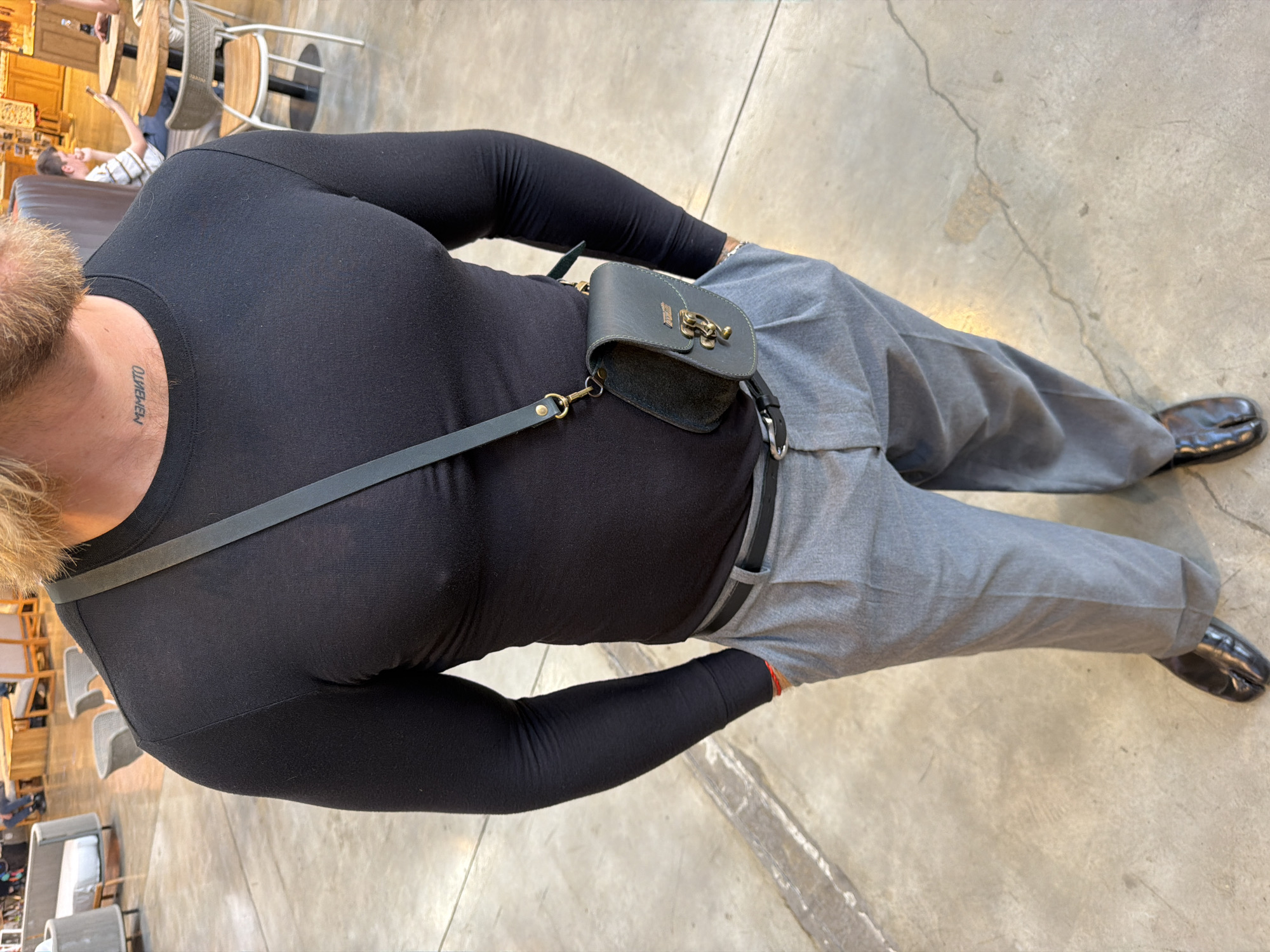 The image shows a person from the chest down, wearing a black long-sleeved shirt and grey pants. The person has a tattoo on their neck that appears to read "MEMENTO". A small, dark grey leather pouch with a metal clasp is attached to a strap that goes across the person's chest. The person is wearing black split-toe shoes. The scene is set indoors, on a concrete floor, with glimpses of outdoor seating areas with wooden tables and chairs visible in the background. The lighting suggests it is daytime. The overall impression is casual and stylish.