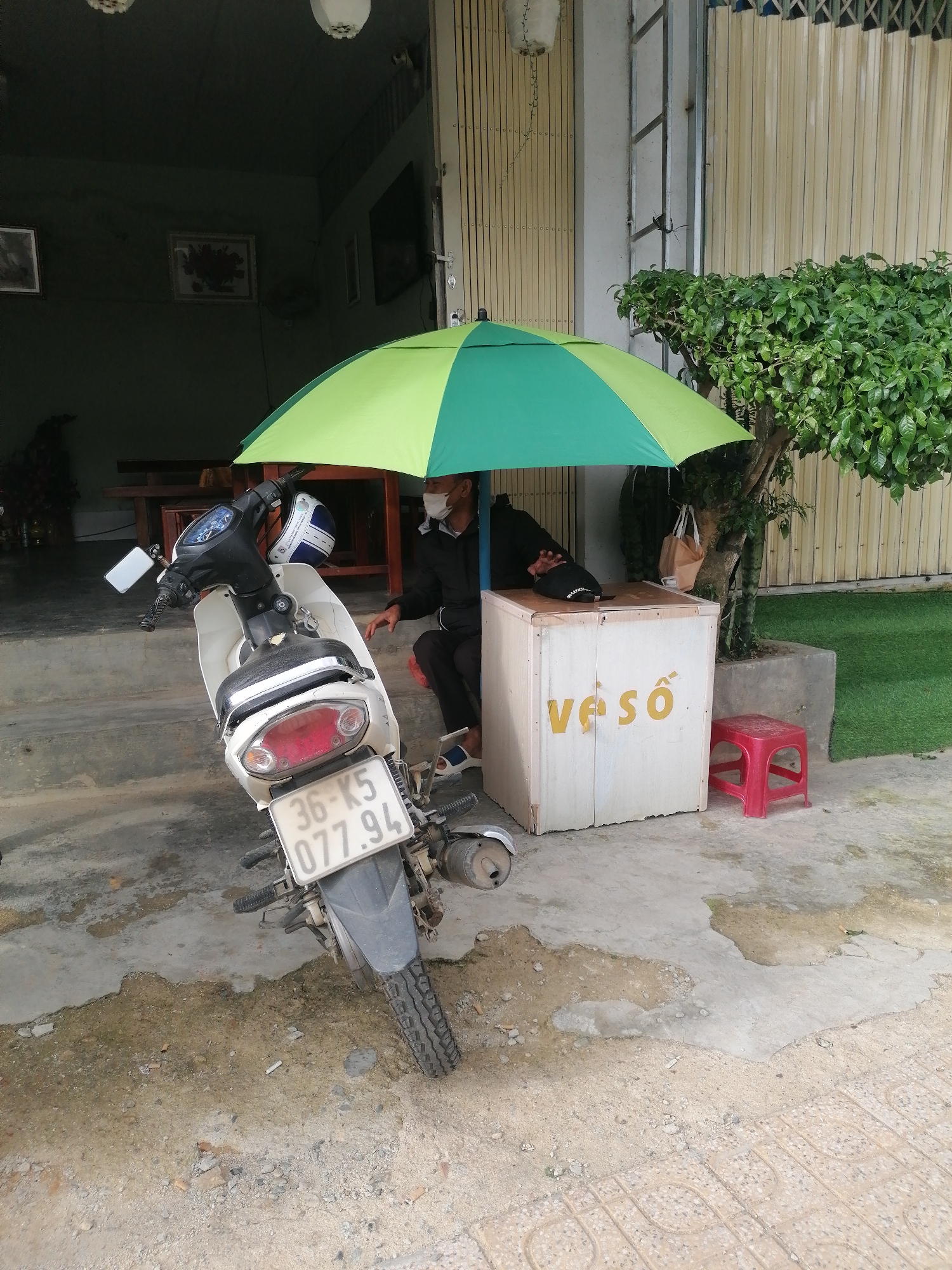 The image depicts a scene outside a building in Xuân Thành, Vietnam. A person wearing a face mask and a black jacket is seated on a red stool under a green and yellow umbrella, next to a white wooden box with yellow lettering. A white motorcycle is parked in front of the umbrella. The ground is paved with concrete and dirt, with some puddles and debris. A tree with green leaves is situated to the right of the wooden box, and a corrugated metal fence is visible in the background. The building appears to be a storefront or shop.