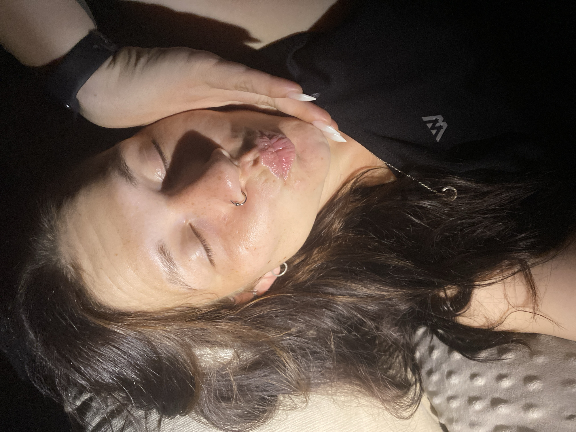 The image shows a close-up, overhead view of a woman with her eyes closed and her lips pursed. Her face is tilted upwards, and a hand is gently cupping her chin, with a fingernail touching her lower lip. She has dark brown hair that falls around her face and shoulders, and she is wearing a silver hoop earring in her left ear, and a nose ring. She is wearing a black shirt with a grey logo that appears to be a stylized "W". The background is dark, with some textured, light-colored fabric visible on the left. There is no discernible indication of a specific location within the city of Chaikovsky, Russia.