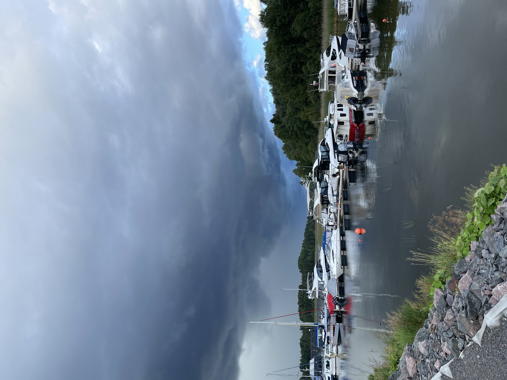 The image shows a marina filled with various boats docked along a pier. The sky is dramatic with dark, heavy clouds gathering, suggesting an impending storm. The water in the marina is calm, reflecting the boats and the sky. Trees line the background of the scene, and a rocky embankment is visible in the foreground. There are no people visible in the image. The location is Berozovo, Russia.
