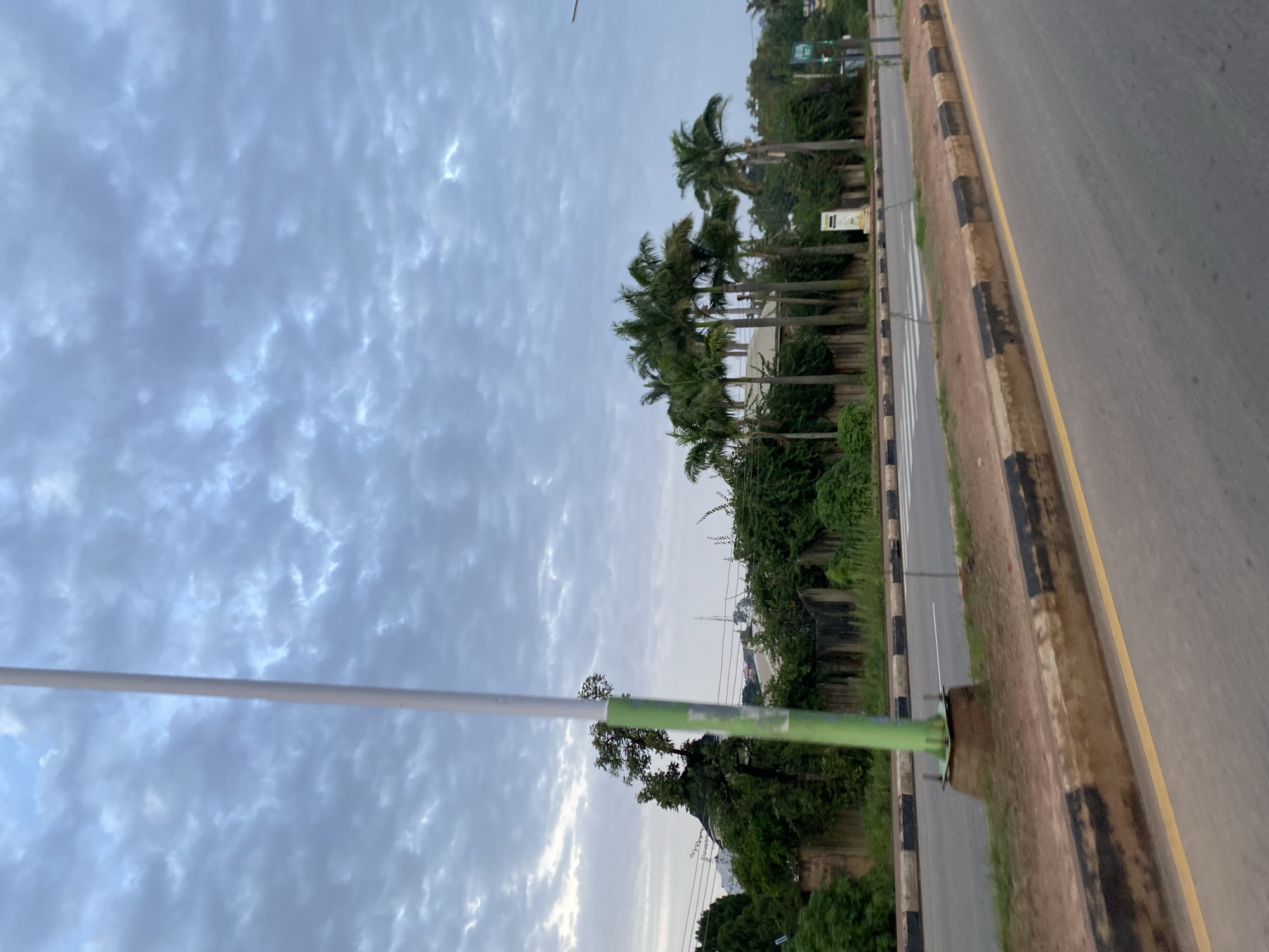 The image depicts a street scene in Jos, Nigeria, under a cloudy sky. In the foreground, a paved road with yellow and black striped curbs runs diagonally from the bottom right to the middle left of the frame. A pedestrian crossing is visible in the middle ground, marked by white stripes. On the left side of the road, a tall, green and white striped pole stands prominently. Behind the road, there is a line of lush green vegetation, including several tall palm trees. A stone wall borders the vegetation, and glimpses of buildings and a telecommunications tower can be seen through the trees in the background. The overall impression is of a moderately urbanized area with significant greenery.