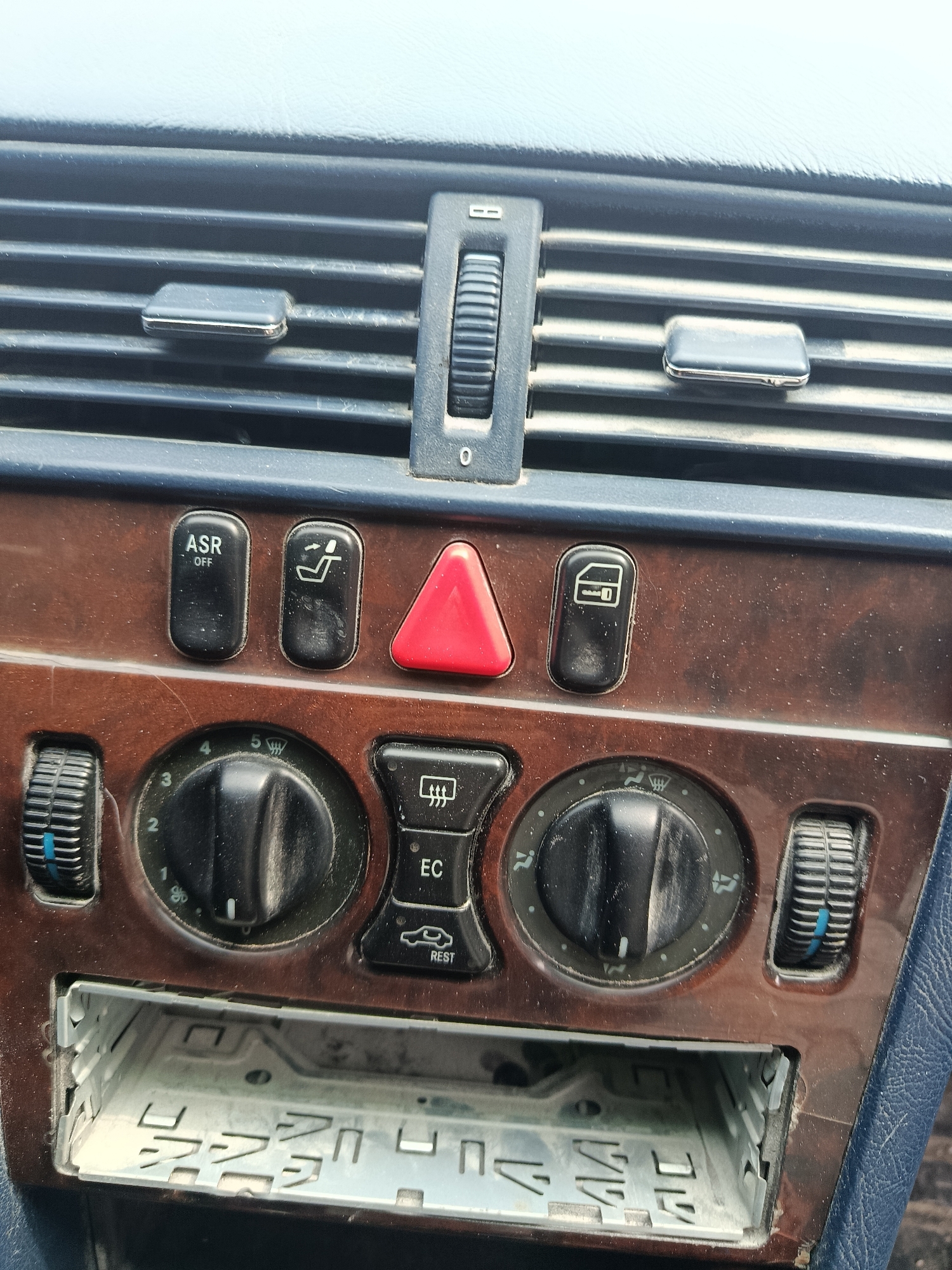 The image shows the dashboard of a car, likely an older model due to its design and style. The dashboard is predominantly brown wood-grain trim with black buttons and knobs, and a blue-gray plastic top section.
Features include:
- Central air vents with adjustable louvers.
- Various buttons with labels such as "ASR OFF", a seat symbol (likely for heated seats), a hazard light button (red triangle), and a window symbol (possibly for window lock).
- Control knobs for fan speed and temperature, with blue accents.
- Buttons labeled "EC" and "REST", likely related to the car's climate control system.
- A rectangular opening where a car radio or entertainment system would typically be located.
The overall condition shows signs of wear and age, with some dust and slight scratches. The setting is inside a car.