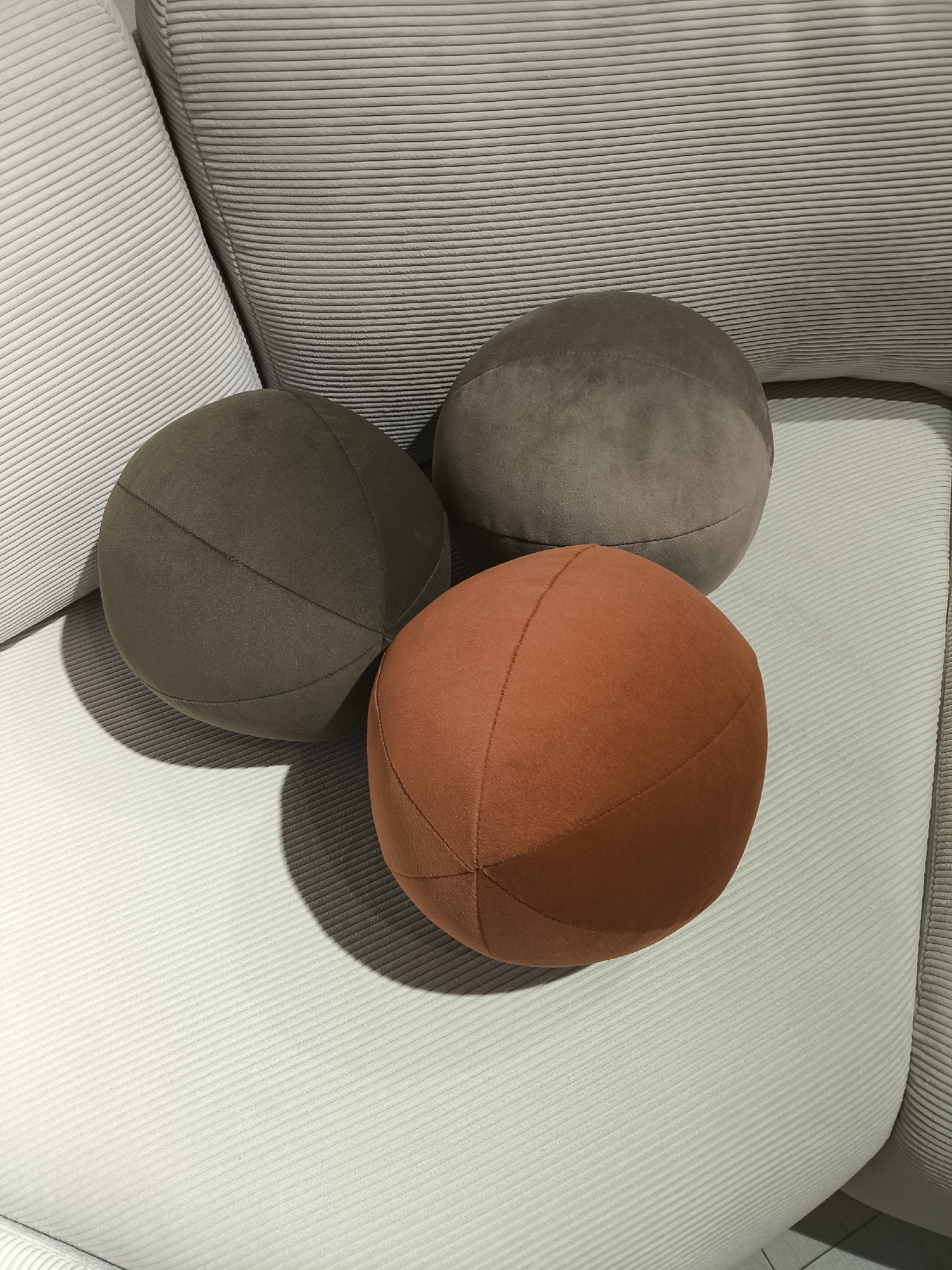 Three spherical pillows are arranged on a light-colored, corduroy-textured sofa. The pillows are brown, gray, and orange. The brown pillow is positioned to the left, the gray pillow is behind and slightly to the right of the brown pillow, and the orange pillow is in the front, partially obscuring the other two. The location is in the city of Khimki, Russia.