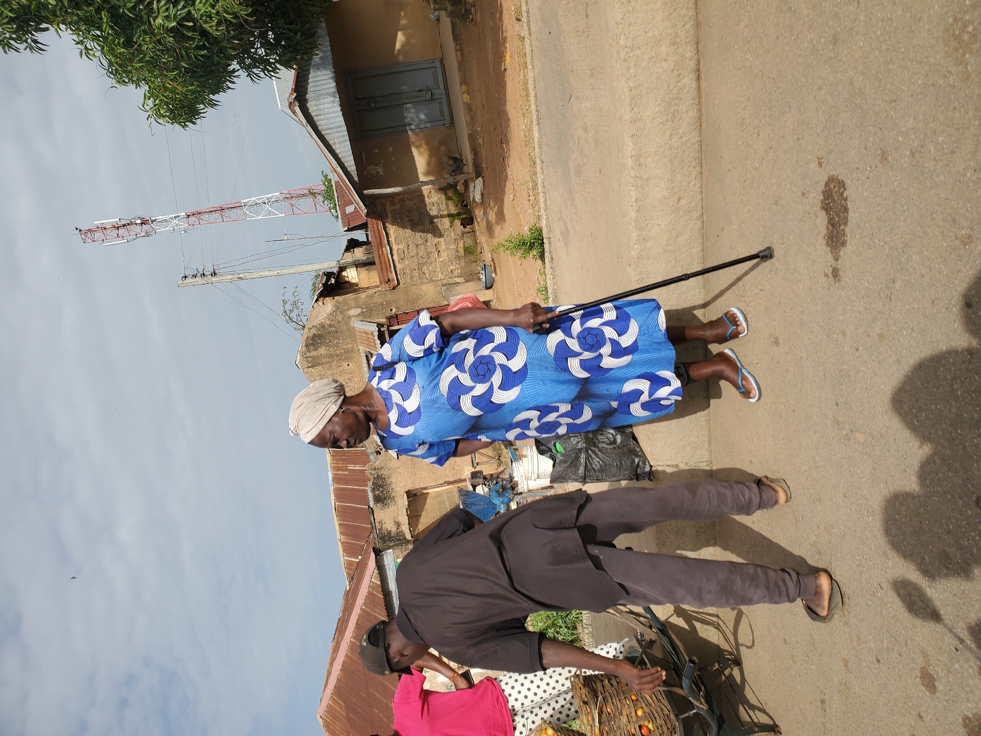In Bukuru, Nigeria, an older woman wearing a light-colored headwrap and a blue and white patterned dress stands on a paved road, leaning on a black cane. She is looking to her right. To her left, a man in a dark t-shirt and grey pants is bending over a cart filled with baskets of produce. A portion of another person in a pink shirt and white polka dot pants is visible to the far left. Behind the people, there are buildings with corrugated metal roofs and some stone construction. A tall, red and white telecommunications tower is visible in the background, along with trees and a cloudy sky.