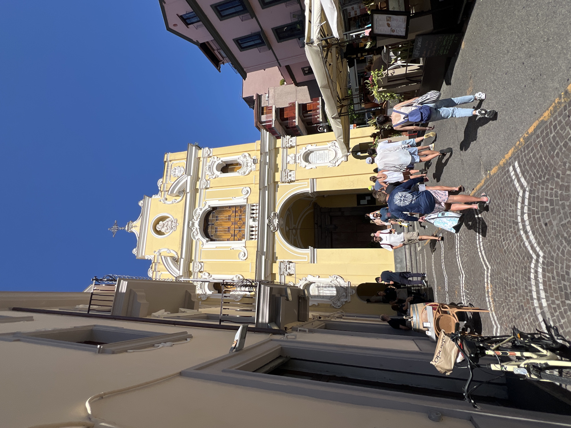The image captures a street scene in Sorrento, Italy, featuring the prominent yellow facade of the Chiesa di San Francesco d'Assisi. The church's ornate Baroque architecture, including its large arched entrance and decorative detailing, is a central element. In the foreground, a cobblestone street with white inlaid patterns leads towards the church. Several people are walking along the street, some heading towards the church and others away from it. They are dressed in casual summer attire. To the right of the street, outdoor seating with umbrellas indicates a cafe or restaurant. A bicycle is parked to the left of the street, near a table and chairs. The sky is a clear, vibrant blue, and the sun is casting shadows, suggesting a bright, daytime setting. The overall impression is one of a lively, picturesque Italian town square.