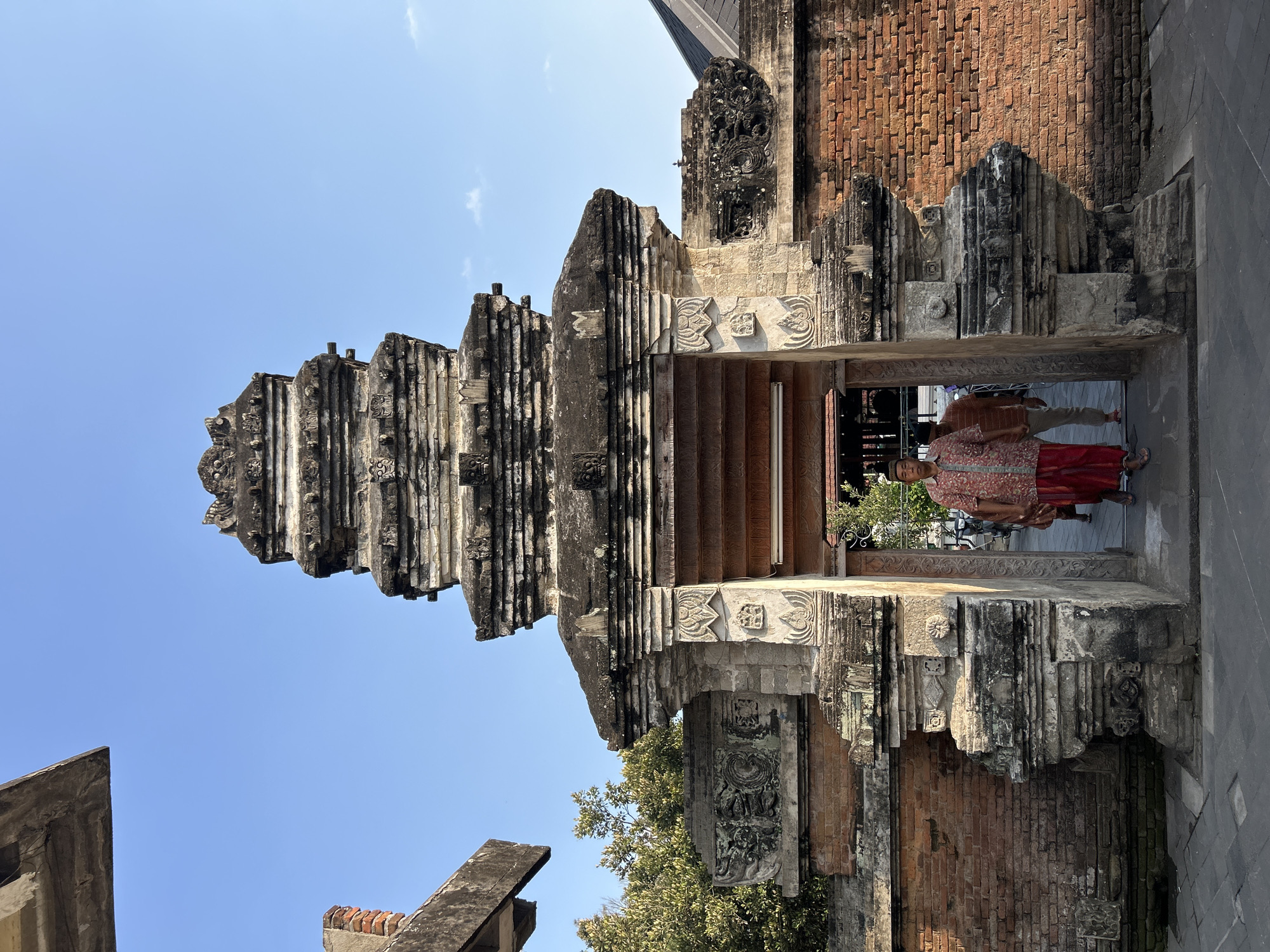 Two men are walking through an ornate stone gateway. The man in the foreground is wearing a red and white patterned shirt and a red sarong. He is walking towards the camera. The man behind him is wearing a brown shirt and white pants, and is walking in the same direction as the foreground man. The gateway is constructed of brick and stone, with intricate carvings. The sky is blue and clear. The scene appears to be the entrance to a temple or mosque in Yogyakarta, Indonesia.