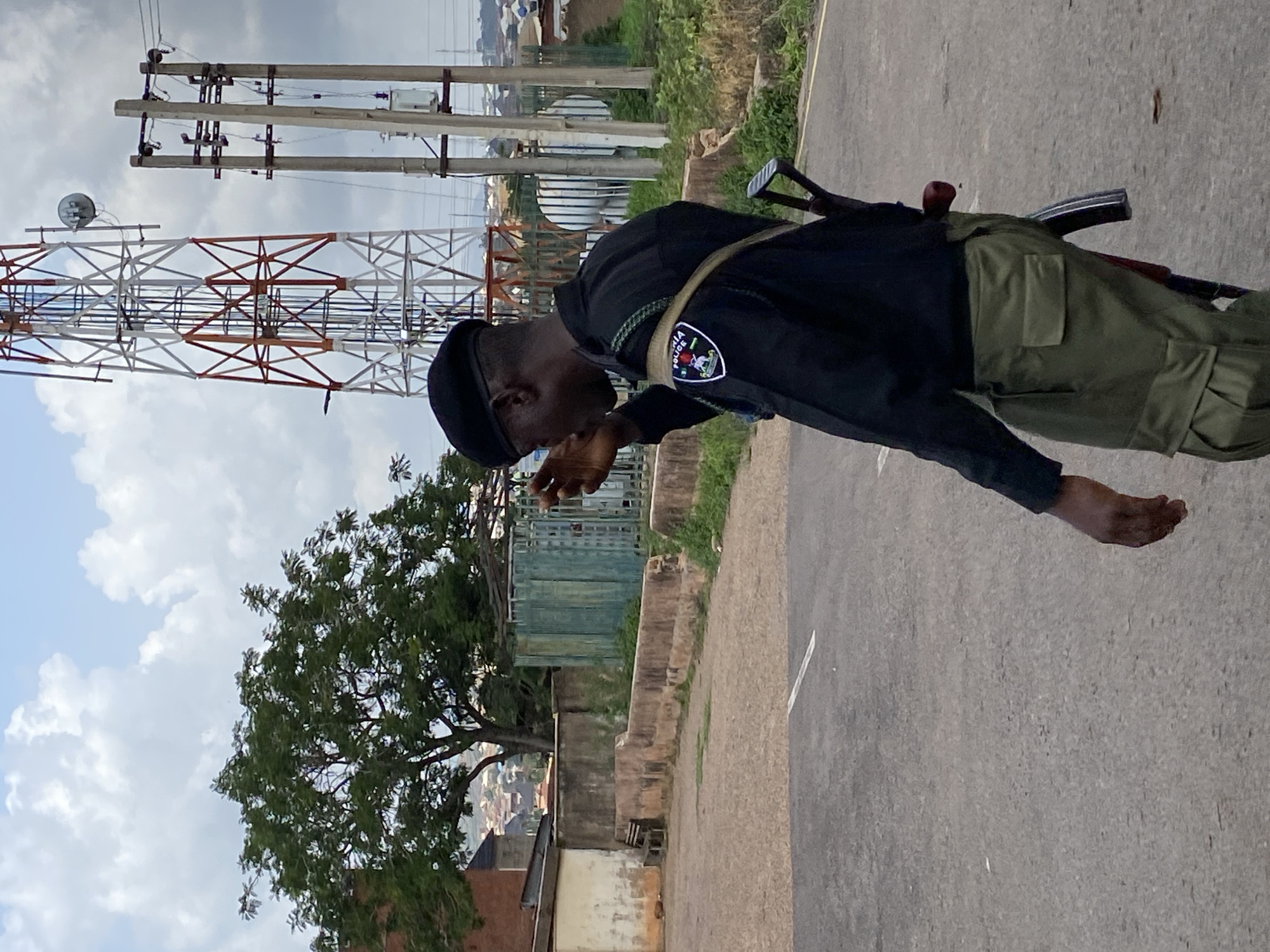 The image shows a person in a black uniform, possibly a police officer, standing outdoors. The person is wearing a black beret and a dark uniform shirt with a patch on the sleeve that reads "NIGERIA POLICE." They are also wearing olive-green pants. The person is holding a rifle that is slung over their shoulder. Their head is bowed and their hand is near their face.
The background includes a large telecommunications tower and a smaller tower with wires and poles. There are also trees, buildings, and a road visible. The sky is cloudy. The image appears to be taken during the day. The presence of the "NIGERIA POLICE" patch and the general appearance of the surroundings suggest the location is in Nigeria. The person's posture might indicate contemplation or fatigue.