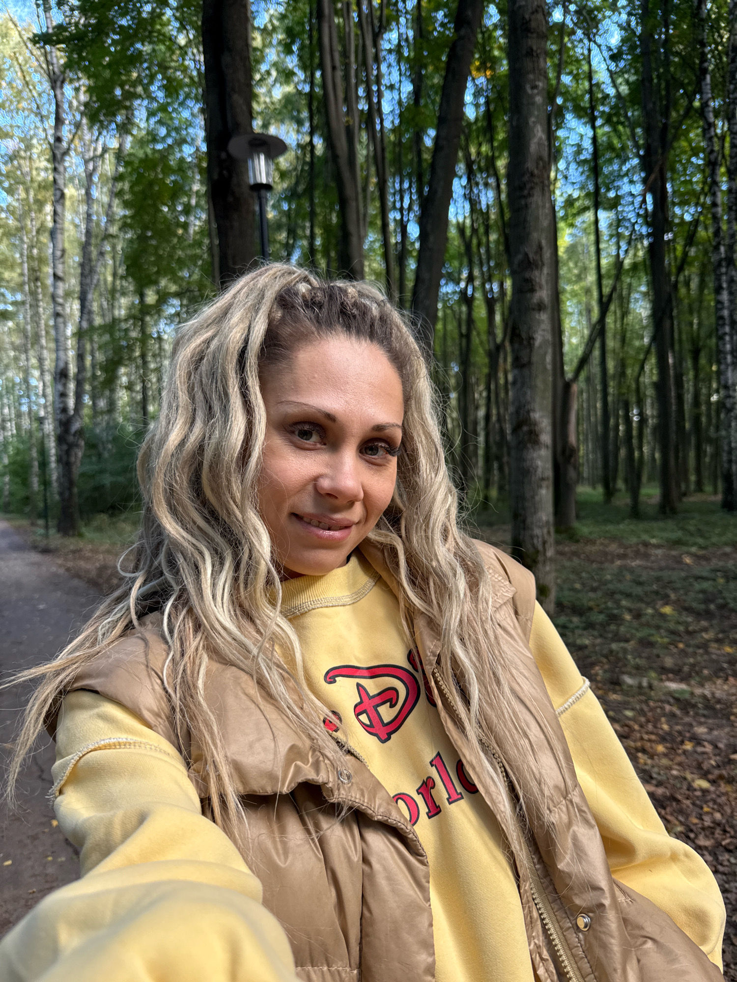 A blonde woman with long, wavy hair is taking a selfie in a park in Rusanovo, Russia. She is wearing a yellow sweatshirt with a red Disney logo and a brown puffer vest. The park has tall trees, fallen leaves, and a paved path. The lighting suggests it is daytime.