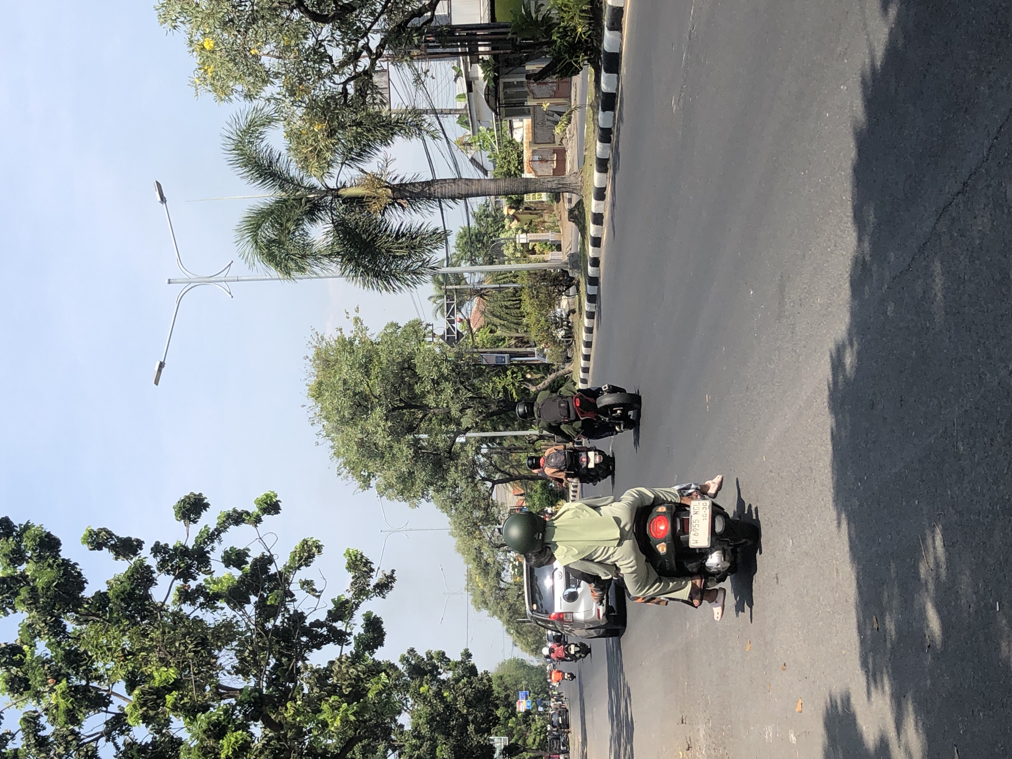 The image depicts a street scene in Larangan, Indonesia, on a sunny day. In the foreground, a person rides on the back of a scooter, wearing a green hijab and long-sleeved shirt, with their feet resting on the scooter's footrest. The scooter has a license plate visible. Ahead of them, a silver car is traveling in the same direction. Further down the road, other individuals on motorcycles are visible, some wearing helmets. The street is paved and appears to be a main road with traffic. Trees line the sides of the road, and there are streetlights and power lines overhead. Buildings and houses are visible in the background, suggesting an urban or suburban setting. The sky is clear and blue. The overall impression is one of everyday traffic on a bright day in Indonesia.