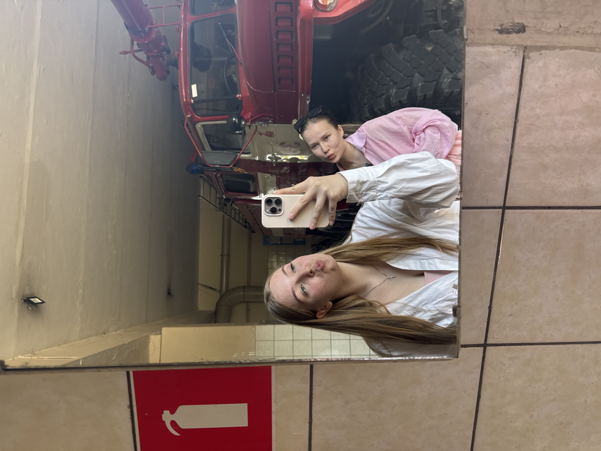 Two women are taking a selfie in a mirror. Behind them, reflected in the mirror, is a red fire truck inside a building. A red fire extinguisher sign is visible on the wall near the bottom of the mirror. The floor is tiled. The location appears to be the fire station in Rusalevka, Russia.