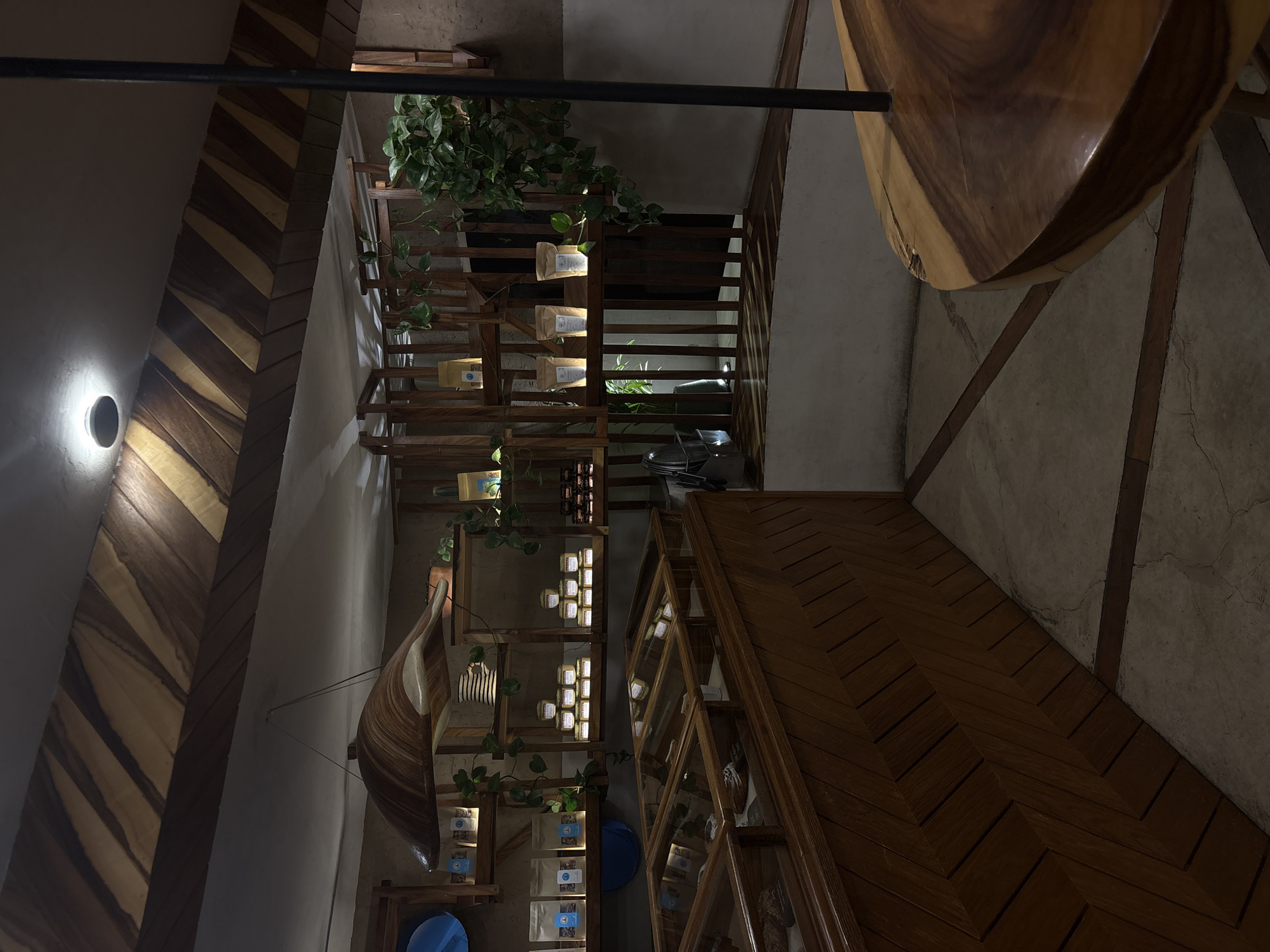 The image depicts an interior space in San Andrés Cholula, Mexico, featuring distinctive wooden design elements. A structural column adorned with a chevron wood pattern is visible to the left, illuminated by a wall-mounted light fixture. A shelving unit, also made of wood, is stocked with bagged items and interspersed with trailing greenery. Adjacent to the shelving unit is a display case with a wood frame, containing a variety of items. Overhead, a wooden table or counter is partially visible. The floor is concrete with patterns. The space appears to be either a retail environment or a uniquely designed café. The lighting is subdued, creating an intimate atmosphere.