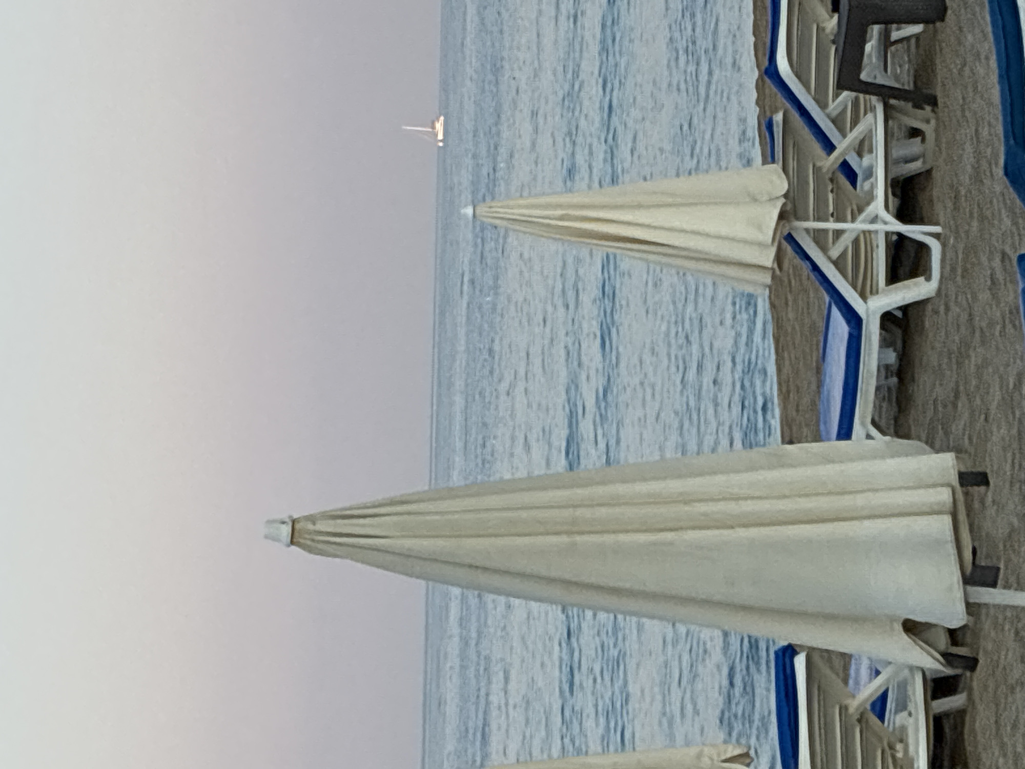 The image captures a serene beach scene in Alanya, Turkey, likely during the early morning or late evening, judging by the soft pastel hues of the sky and the gentle calm of the sea. In the foreground, several closed beach umbrellas stand tall on the sandy shore, their cream-colored fabric neatly folded. Two of these umbrellas are particularly prominent, with one on the left occupying a significant portion of the frame, and another smaller one to its right.
Adjacent to the umbrellas are rows of empty white sun loungers with blue cushions, arranged on the sand, suggesting a quiet moment before or after a busy day at the beach. The loungers are unoccupied, reinforcing the sense of tranquility.
The sea stretches out beyond the beach, its surface a tranquil expanse of blue-grey water, with subtle ripples indicating a light breeze. In the far distance, a small white sailboat with its sails up is visible on the horizon, adding a touch of maritime charm to the seascape. The overall atmosphere is one of peaceful repose, characteristic of Alanya's beautiful coastline.