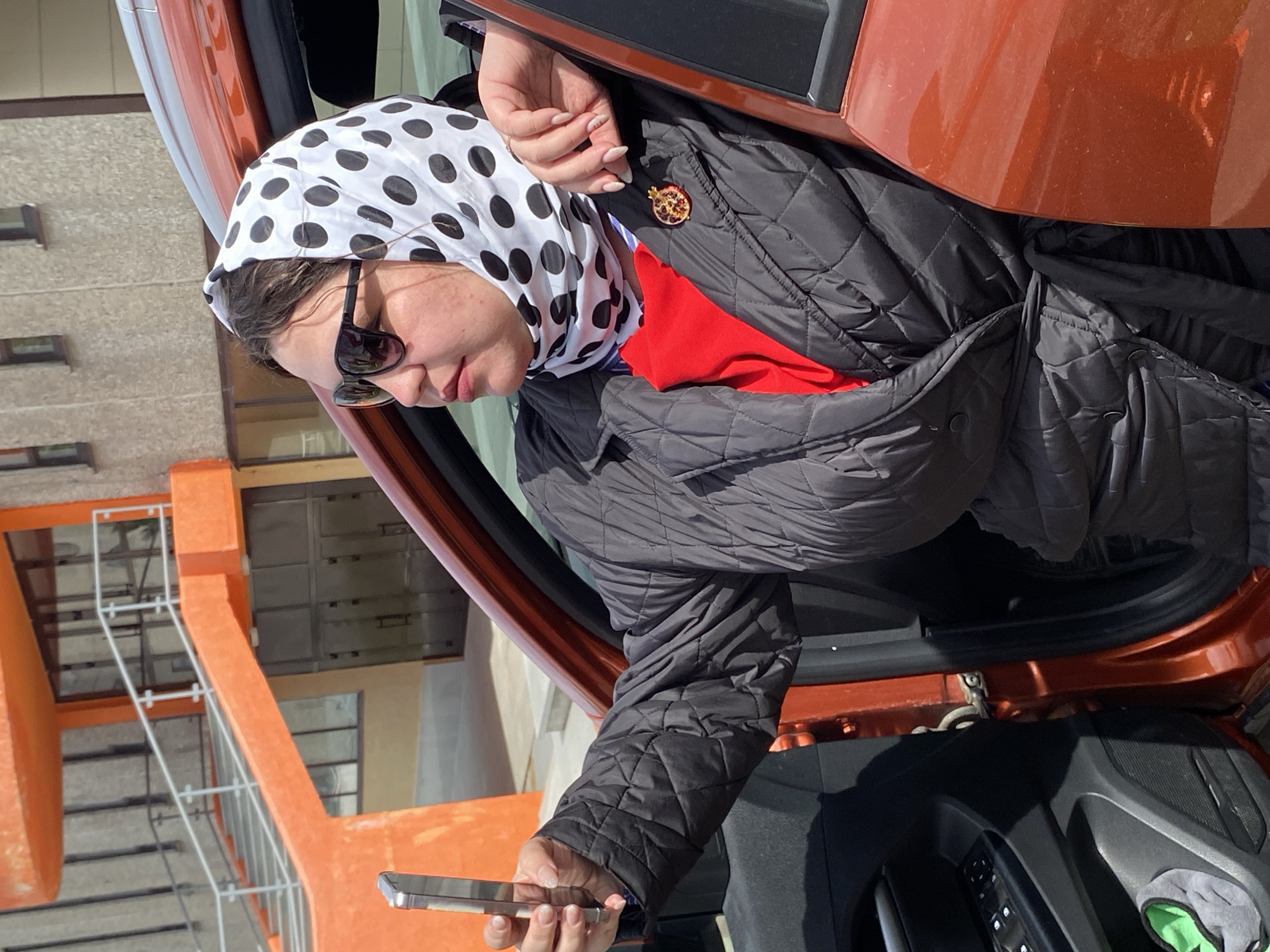 A woman is depicted leaning out of an open car door on a bright, sunny day in Markovskiy, Russia. She wears a white headscarf adorned with black polka dots, dark sunglasses, and a black quilted jacket over a red shirt. A distinctive brooch, shaped like a pomegranate with red and gold details, is pinned to her jacket. Her right hand, with light-colored fingernail polish, firmly grasps a silver smartphone.
The car's exterior, visible around the doorframe, is a warm orange or burnt sienna color. The interior of the car door, including controls and part of the speaker grille, is also visible. In the background, a light-colored building with multiple dark-framed windows stands under clear skies. Adjacent to this building, an prominent orange architectural structure with white railings, possibly an outdoor staircase or a part of another building, ascends into the frame. The overall scene suggests an urban or suburban setting. The woman's expression, partially hidden by her sunglasses, appears neutral and focused as she holds her phone.