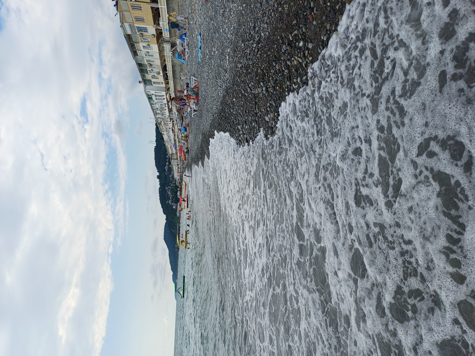 The image depicts a busy beach scene in Sochi, Russia. The perspective is from the water's edge, looking towards the shore. In the foreground, foamy waves are rolling in, obscuring the pebbles of the beach. The beach itself is lined with numerous people relaxing on loungers and towels, their forms dotted amongst the dark grey and multicolored pebbles. Behind the beach, a long, multi-story building with balconies and many windows stretches along the coastline, suggesting a resort or hotel. Further in the distance, a steep, tree-covered mountainside rises, with some buildings visible on its slopes.
The sky is partly cloudy, with patches of blue peeking through soft white clouds, indicating fair weather. The time of day appears to be daytime, likely late morning or afternoon, given the lighting. People are engaged in typical beach activities such as sunbathing and lounging. A few colorful umbrellas and beach accessories are visible. In the water, there are some inflatable toys and a paddleboat, suggesting recreational activities on the water. The overall atmosphere seems relaxed and summery, with a good number of people enjoying the seaside. No visible text is present in the image.