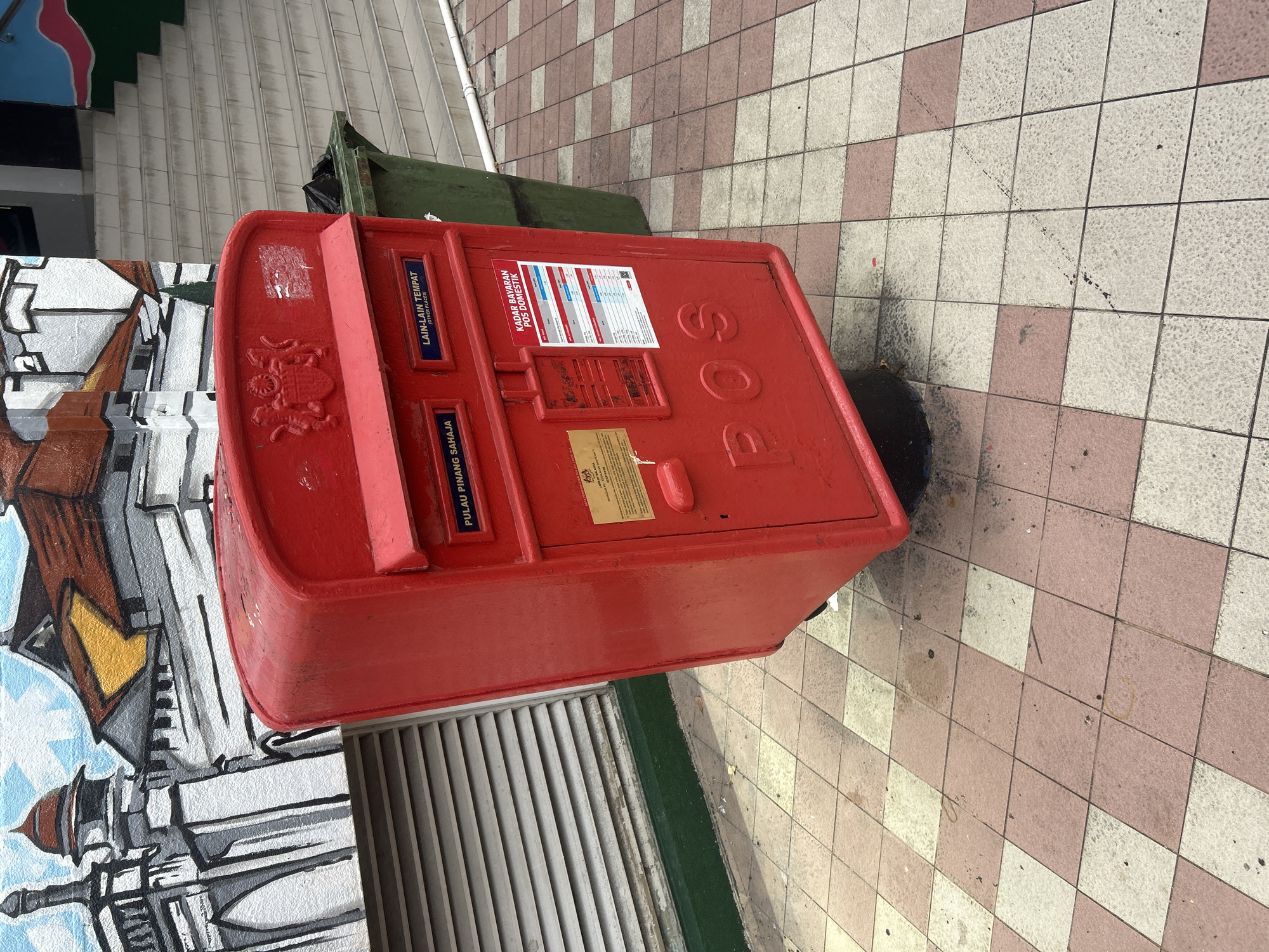 A bright red, vintage-style post box stands tilted on a patterned tiled sidewalk in George Town, Malaysia. The post box, made of metal, features a royal crest on its upper front, and has two slots for mail. The left slot is labeled "PULAU PINANG SAHAJA" (Penang Only), and the right slot is labeled "LAIN-LAIN TEMPAT" (Other Places). Below the slots, a colorful chart detailing "KADAR BAYARAN POS DOMESTIK" (Domestic Postal Rates) is affixed. The word "POS" is embossed in large, stylized letters on the lower front of the box. To the left of the post box is a grey, slatted ventilation unit, and to the right, a green trash receptacle is visible near a set of concrete stairs. The background features a mural depicting a mosque or temple-like building with a blue sky. The sidewalk is tiled with alternating cream and reddish-pink squares. The scene appears to be outdoors in a public area.