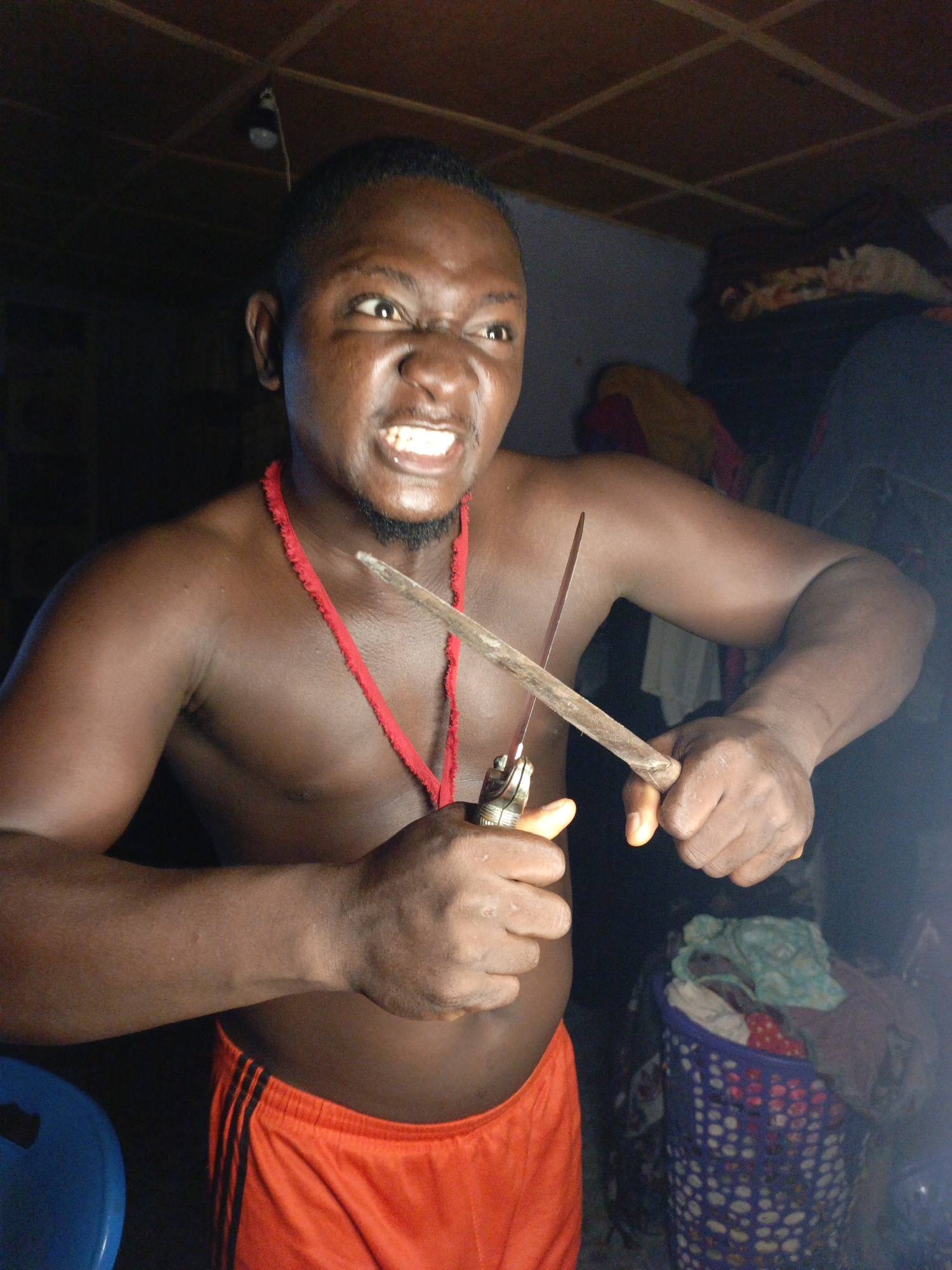 In an interior scene, a shirtless Black man with a red necklace around his neck and wearing orange shorts glares at the camera, holding two knives. One knife is held in his right hand, its blade pointed upwards. The other knife is held in his left hand, its blade pointed downwards and forwards. The man’s face is contorted in a grimace, with his teeth bared and eyes narrowed. His arms are muscular, and his chest and abdomen are visible. In the background, clothing and a laundry basket are visible, suggesting a domestic environment. The lighting is dim, with a single light bulb hanging from the ceiling. The man's posture and expression suggest aggression or a ritualistic act.