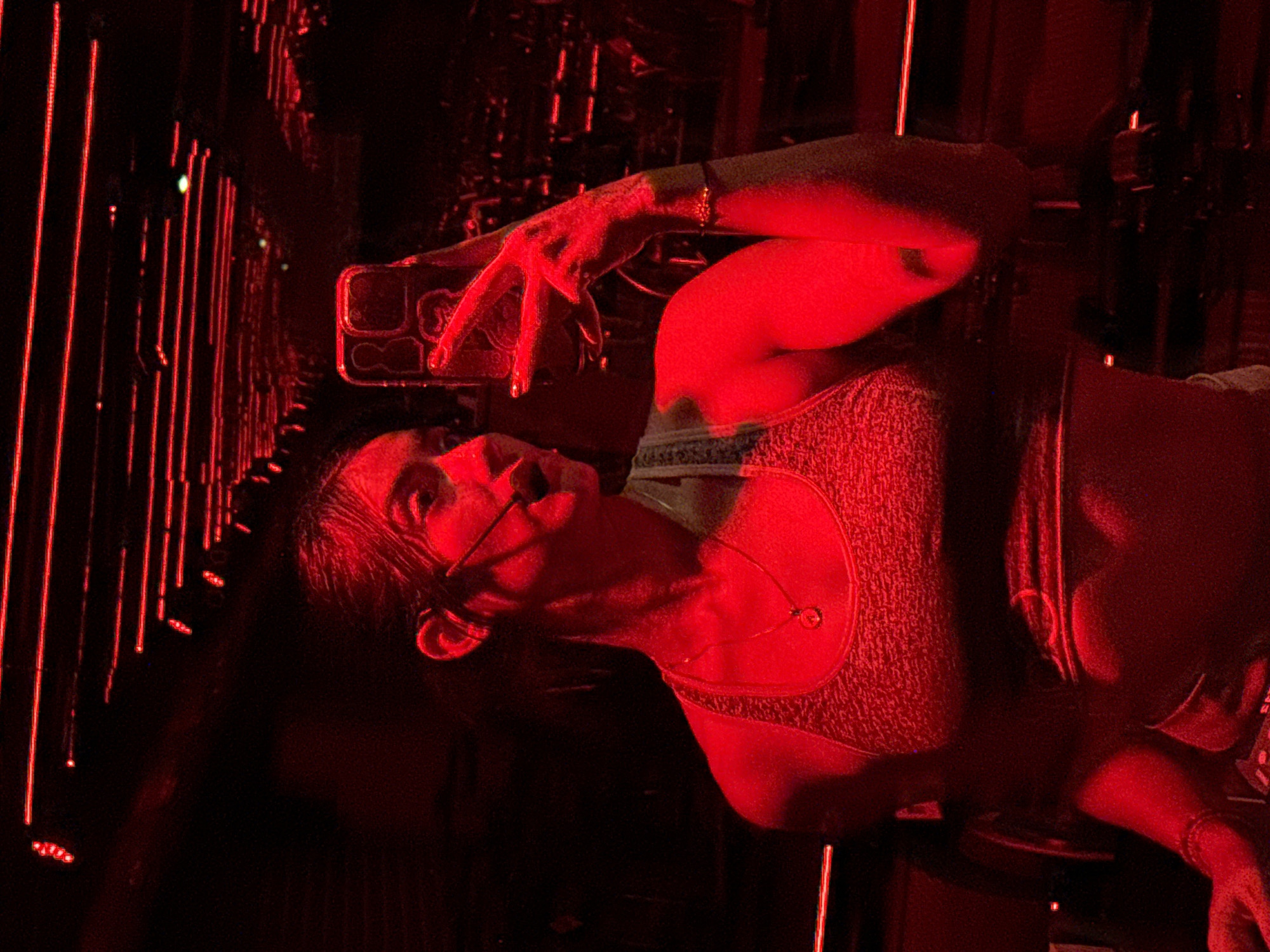 A woman is seen taking a selfie in a mirrored room with red lighting. The room appears to be a nightclub or bar in the city of Puebla, Mexico. The room contains linear lights and mirrored walls. The woman is wearing a light-colored, possibly silver, tank top and jewelry. She is holding a smartphone in her left hand, with her fingers posed in front of the camera. The woman is facing the camera at an angle, with her head turned slightly to the side. Her hair is dark. There is a microphone.