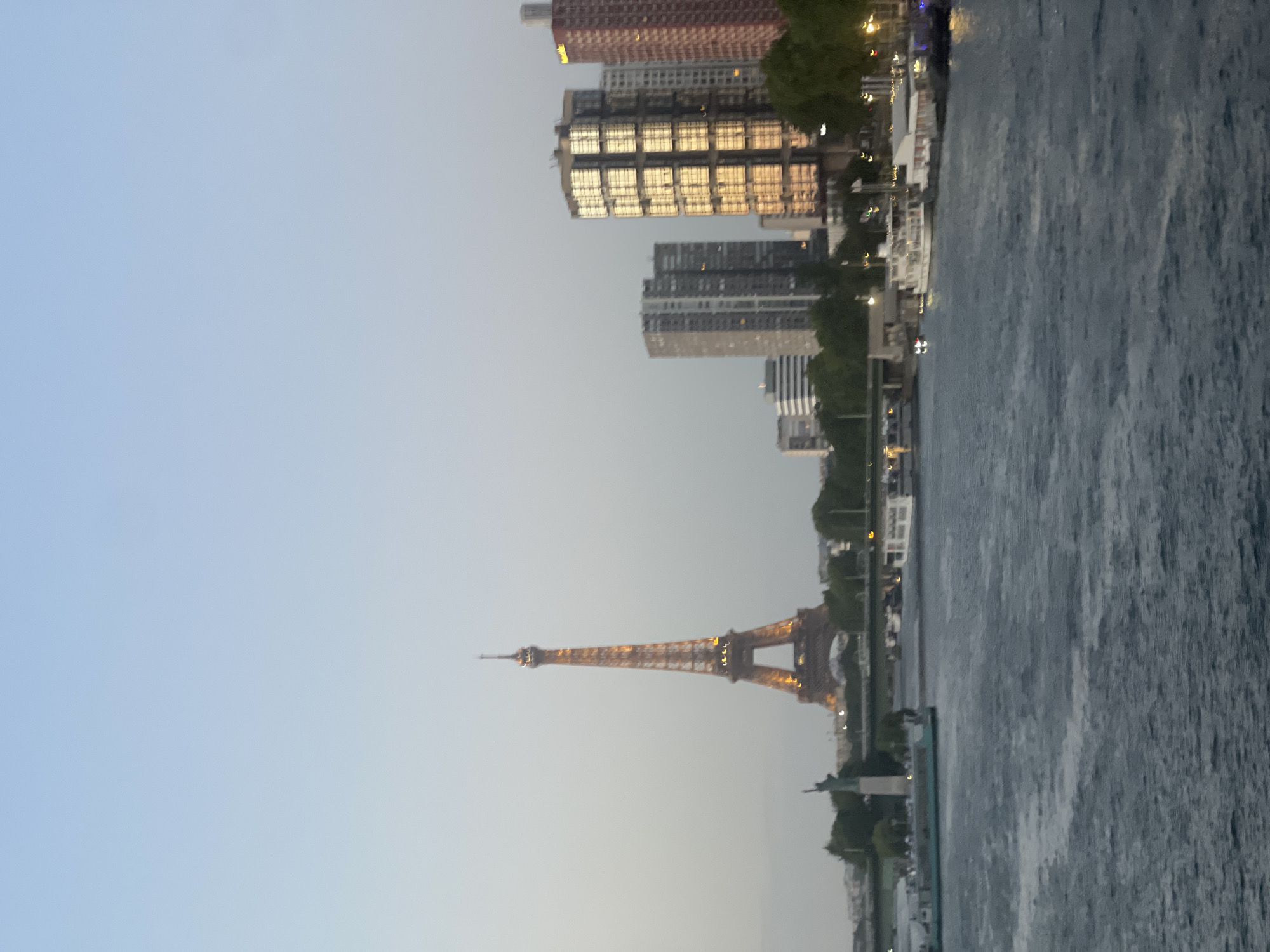 The image shows a view of the Eiffel Tower in Paris, France, at dusk. The Eiffel Tower is illuminated with golden lights and stands prominently in the center of the frame. To the left of the Eiffel Tower, a smaller replica of the Statue of Liberty is visible. In the foreground, the Seine River flows, with several boats docked along the bank. Modern buildings with reflective windows are visible on the right side of the image, contrasting with the historic landmark. The sky is a soft gradient of blue and gray, indicating the transition from day to night. Trees line the riverbank, and a bridge can be seen in the background. The overall scene captures a quintessential Parisian cityscape.