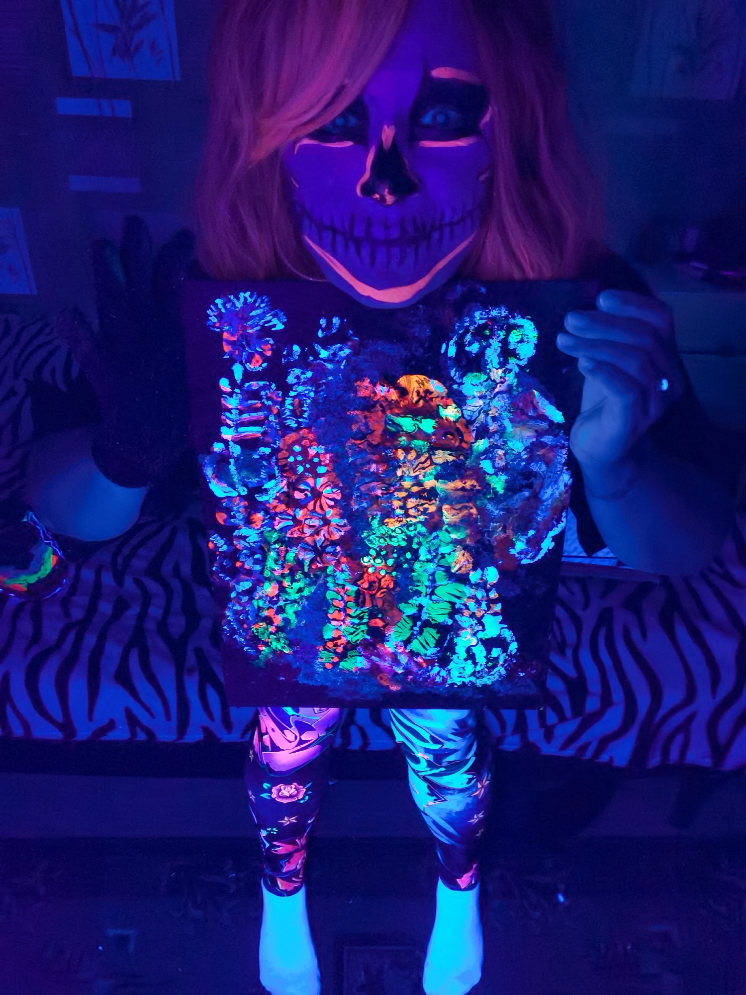 A person is depicted in a blacklight-illuminated indoor setting, showcasing a piece of art they are holding. The individual, whose gender is not explicitly clear but appears to be female with long hair, wears glow-in-the-dark skull face paint in shades of bright orange and purple. Their hair, possibly a wig, is a vibrant glowing orange.
They are dressed in dark clothing, including leggings adorned with patterns like stars and roses that glow in various colors (pink, blue, purple, orange). Their socks are also glowing white. The person holds up a square canvas or board covered in a dense, abstract array of luminous paint. The artwork features intricate, organic shapes resembling coral, flowers, or microbial structures, glowing vividly in blues, greens, oranges, and purples.
Behind the person, a zebra-print patterned surface (likely bedding or furniture) is visible, with its white stripes glowing faintly blue under the blacklight. To the left, on this surface, a small bowl or cup containing a luminous, multi-colored liquid or paint is present. In the background, on a dark wall, several small rectangular canvases or pictures are subtly glowing blue.
The scene is set indoors, likely at night or in a darkened room, making optimal use of the blacklight to highlight the fluorescent paints and materials. The activity appears to be an artistic display or a creative session, possibly with a festive or Halloween theme suggested by the skull makeup. The person looks directly at the viewer. There is no visible text in the image. The location context provided is Chaikovsky, Russia, suggesting this activity is taking place there.