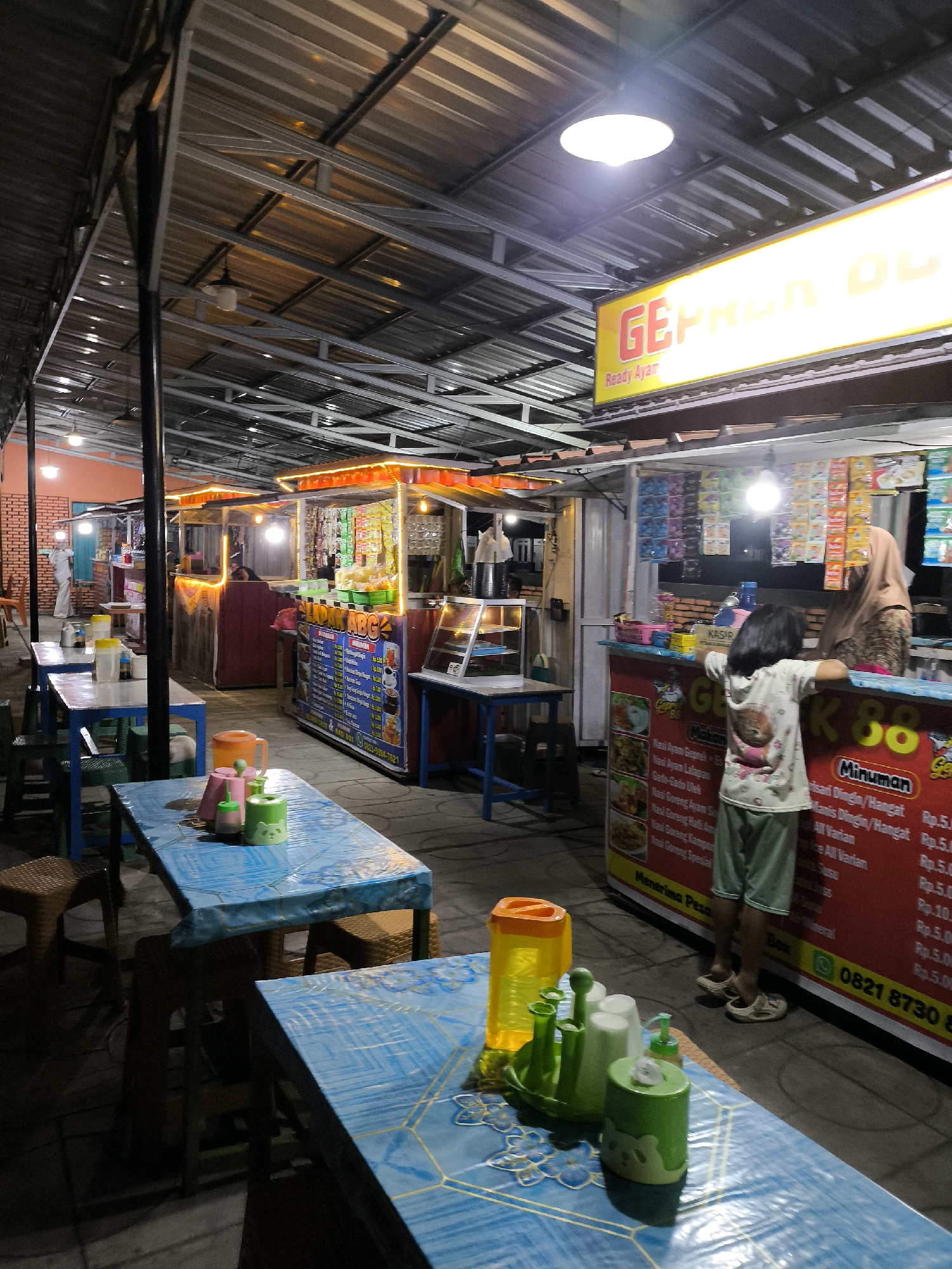 An exterior, eye-level, medium shot captures a food stall area at night. The stalls are covered by a metal roof supported by metal beams, with incandescent lights illuminating the scene. In the foreground, a blue table covered with a plastic tablecloth is set with green condiment containers and a yellow plastic jug. To the right of the table, a young girl wearing a t-shirt with a cat print and green shorts stands facing a food stall. She leans on the counter, looking towards the stall where a woman wearing a hijab and an apron is partially visible. The stall displays menus and various items for sale. Another stall to the left features a similar setup with tables and chairs. The overall environment suggests a casual dining space in a city like Hayahaya, Indonesia, where street food vendors commonly operate.