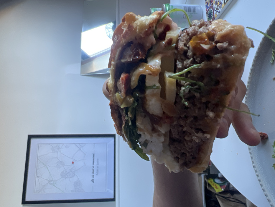 The image depicts an indoor scene, likely a residence in Carnin, France. A large, multi-layered burger, partially consumed, is held by a light-skinned hand in the foreground. The burger features a brioche-style bun, multiple thick ground beef patties, distinct layers of white melted cheese, yellow cheese slices, green leafy vegetables (appearing to be arugula), strips of cooked bacon, caramelized onions, and drizzles of a brown-orange sauce. A noticeable bite has been taken from the burger's front, and small food particles are visible on a light-colored, textured plate below.
In the background, against a plain white wall, a rectangular black-framed map hangs in the upper left quadrant. The map is white with grey street or road lines and features a small red heart marking a specific point. Below the map, French text reads "Là où tout a commencé..." (Where it all began...) and "Aden et Julia." To the right of the map, on a white surface, a rectangular mirror is positioned, reflecting a bright window or light source, showing parts of what appears to be a curtain or blinds and an external architectural element. Further to the right on the white surface, two green beverage cans are stacked, and a small object with a colorful, geometric, possibly tiled pattern is visible. A crumpled yellow plastic bag is partially visible in the lower left corner behind the plate. The overall lighting is bright and diffused.