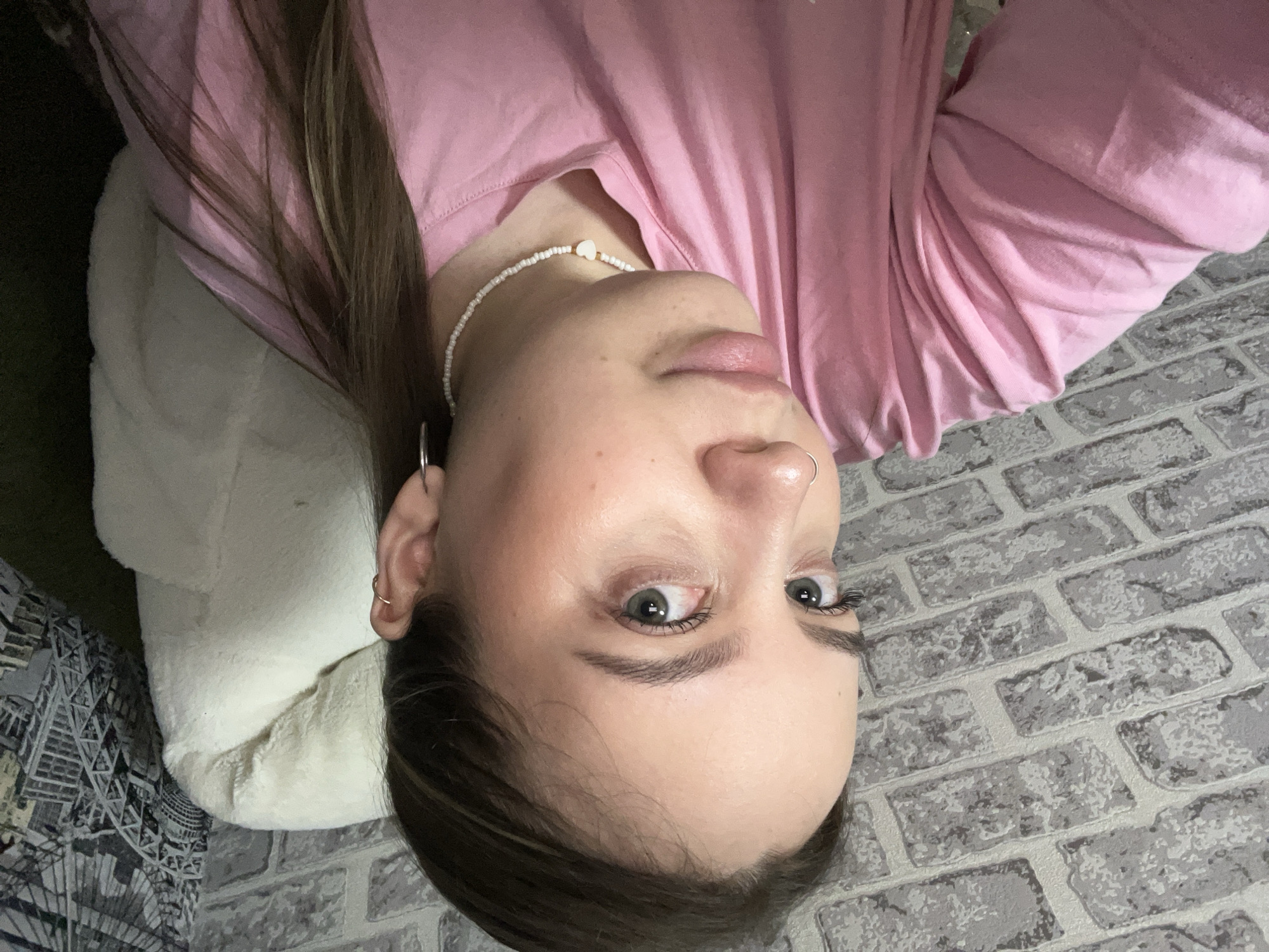 A woman with long brown hair is angled with her head toward the bottom of the frame and her body towards the top. She is wearing a pink long-sleeved shirt and a white beaded necklace. She has a nose ring and earrings. She is resting on a white pillow with a patterned grey and black fabric visible on the left. A gray brick wall is behind her. The location is not discernible from the image.
