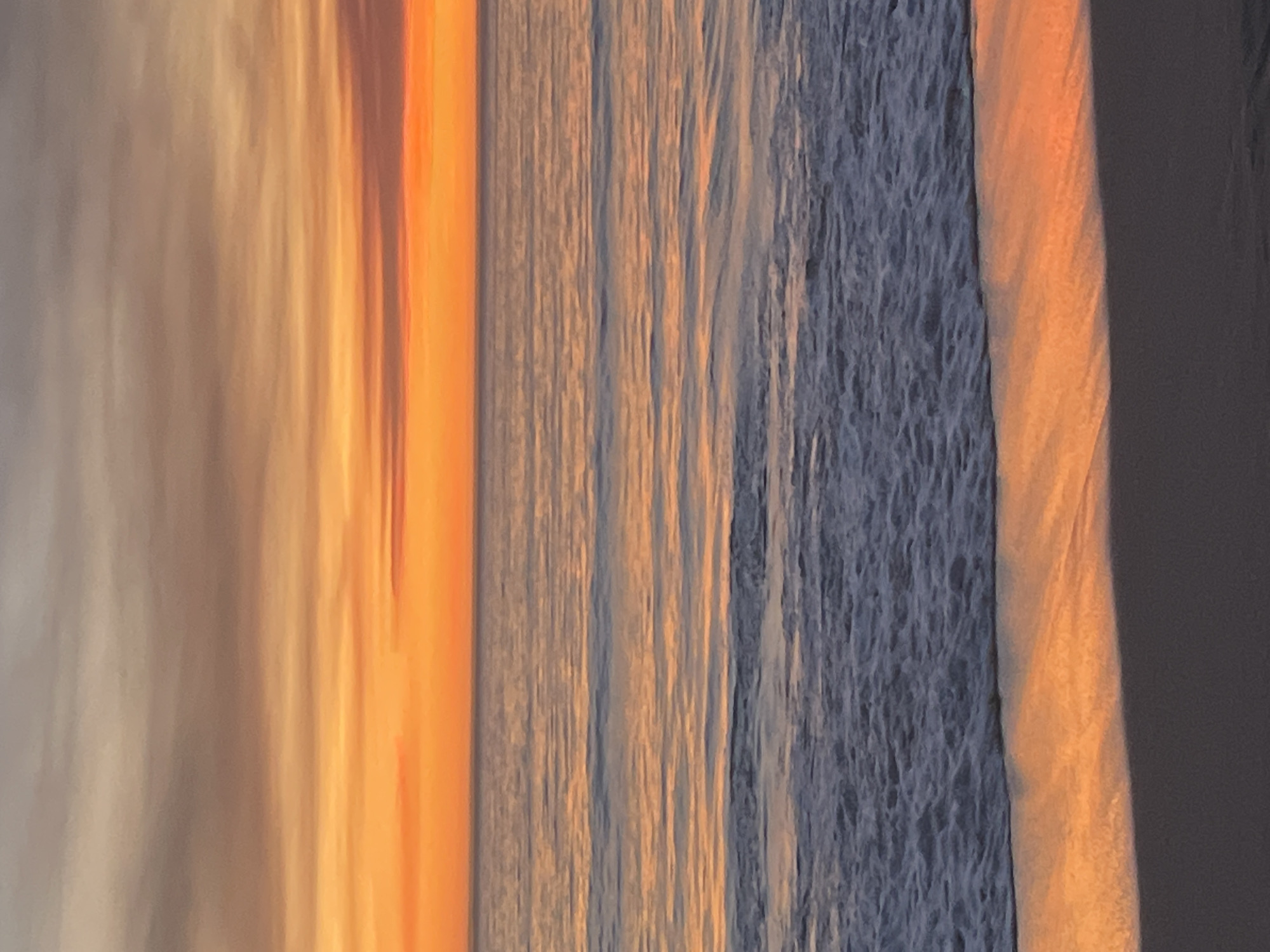 The image depicts a serene beach scene at sunset, with the sky a vibrant canvas of orange and purple hues. The ocean stretches out to the horizon, reflecting the warm colors of the sky. Gentle waves lap at the shore, creating a foamy white edge against the dark sand. This photograph was taken at Praia de Riazor, a popular urban beach located in the city of A Coruña, Spain. No people or specific objects are visible in the frame, emphasizing the natural beauty of the coastal landscape.