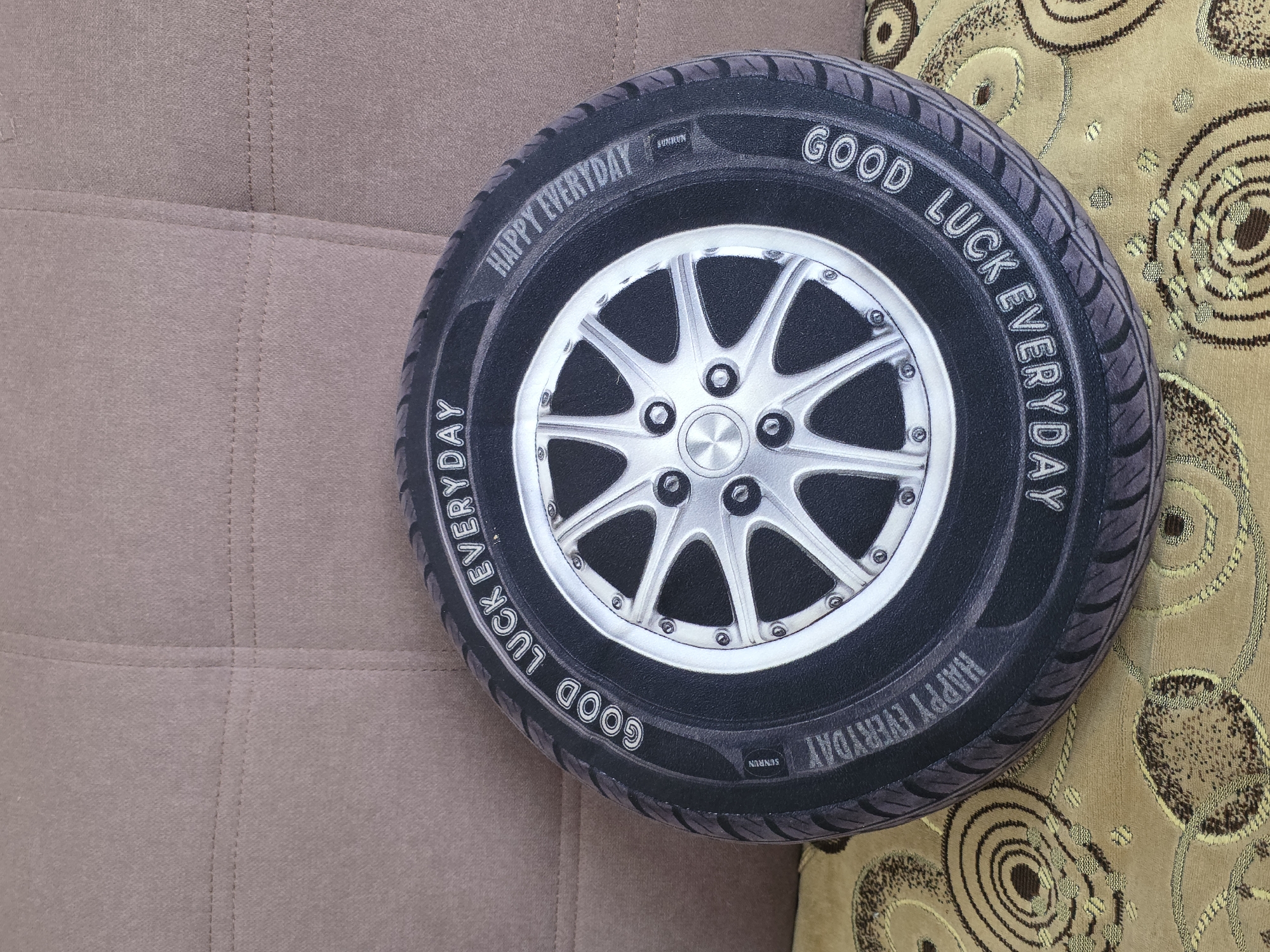 A circular tire-shaped pillow with a silver rim design is placed on a beige, textured surface, likely a couch. The pillow has text printed on its outer edge, reading "GOOD LUCK EVERYDAY" in one direction and "HAPPY EVERYDAY" in the other. The design of the tire includes tread patterns. The background surface is a muted brown color with horizontal stitching creating a panel effect. The pillow rests on a patterned fabric with concentric circles in shades of brown and gold. No people or actions are depicted. The scene is static and indoors. The image does not provide any information about the city of Qarabalıq, Kazakhstan.