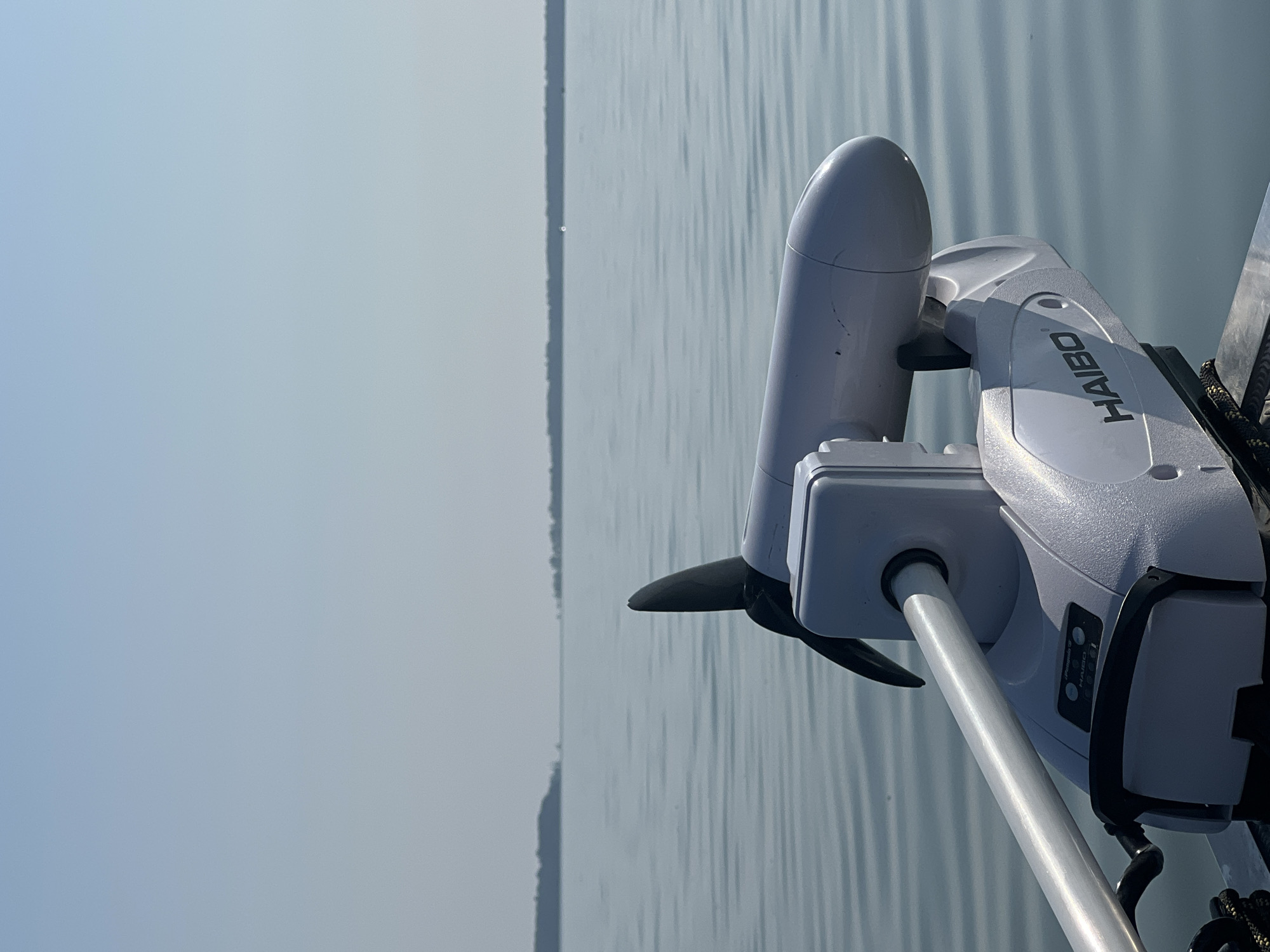 A close-up, eye-level shot captures a white electric trolling motor on a boat on a calm body of water. The motor is the primary subject, with its propeller, shaft, and casing clearly visible. The brand name "HAIBO" is emblazoned on the side of the motor. Beyond the boat, the water stretches to the horizon, meeting a hazy sky. A distant, tree-lined shoreline is visible in the background. The scene is serene and appears to be a sunny day. The location is identified as Beregovoe, Russia, a coastal settlement.