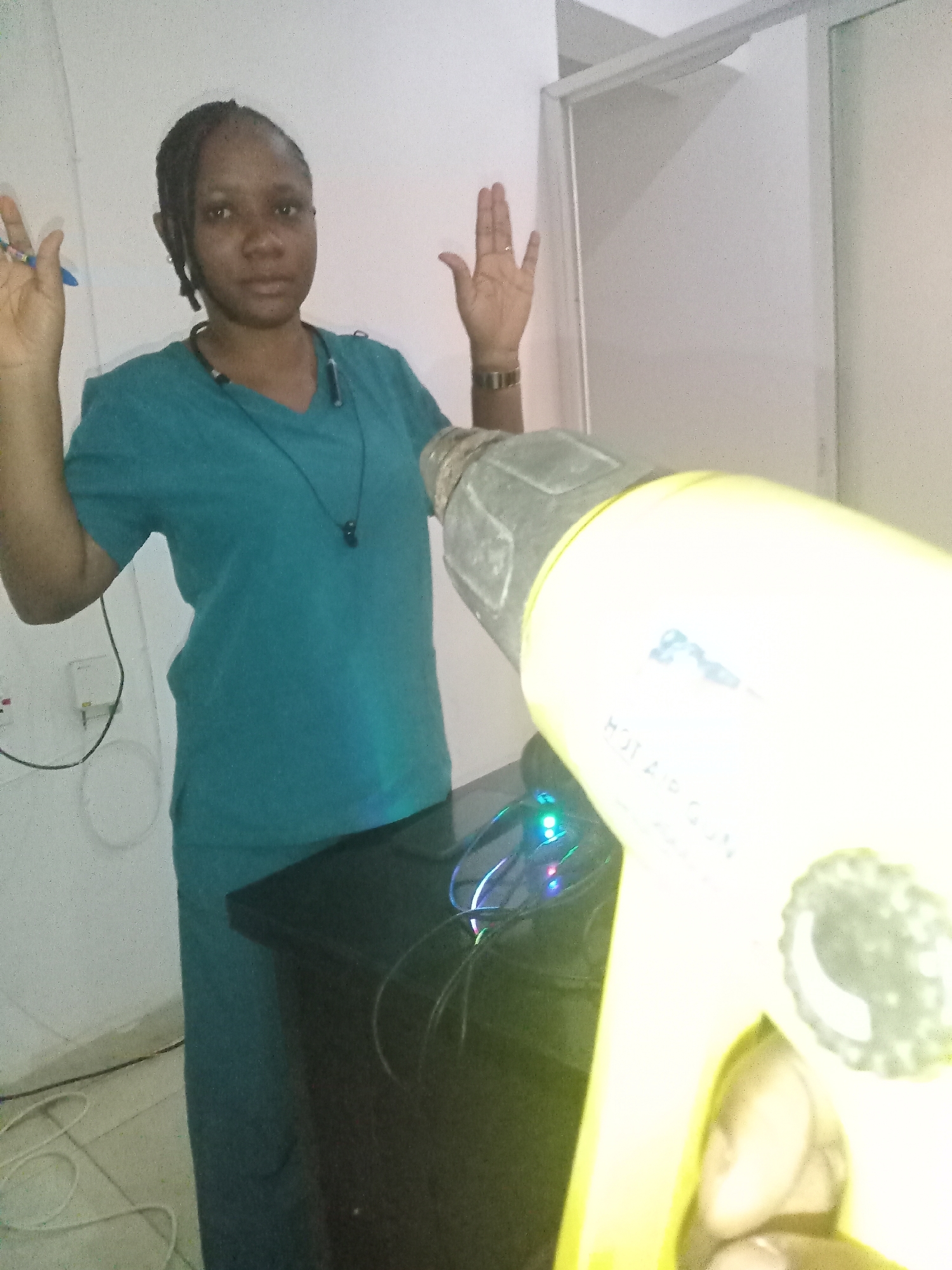 A woman in a teal scrub suit stands with both hands raised near a black cabinet in a room in Gwarinpa, Nigeria. A yellow hot air gun is in the foreground, partially obscuring the cabinet. A power outlet and cord are visible on the wall behind the woman. A glass door or window frame is to the right. The lighting is bright and somewhat harsh.