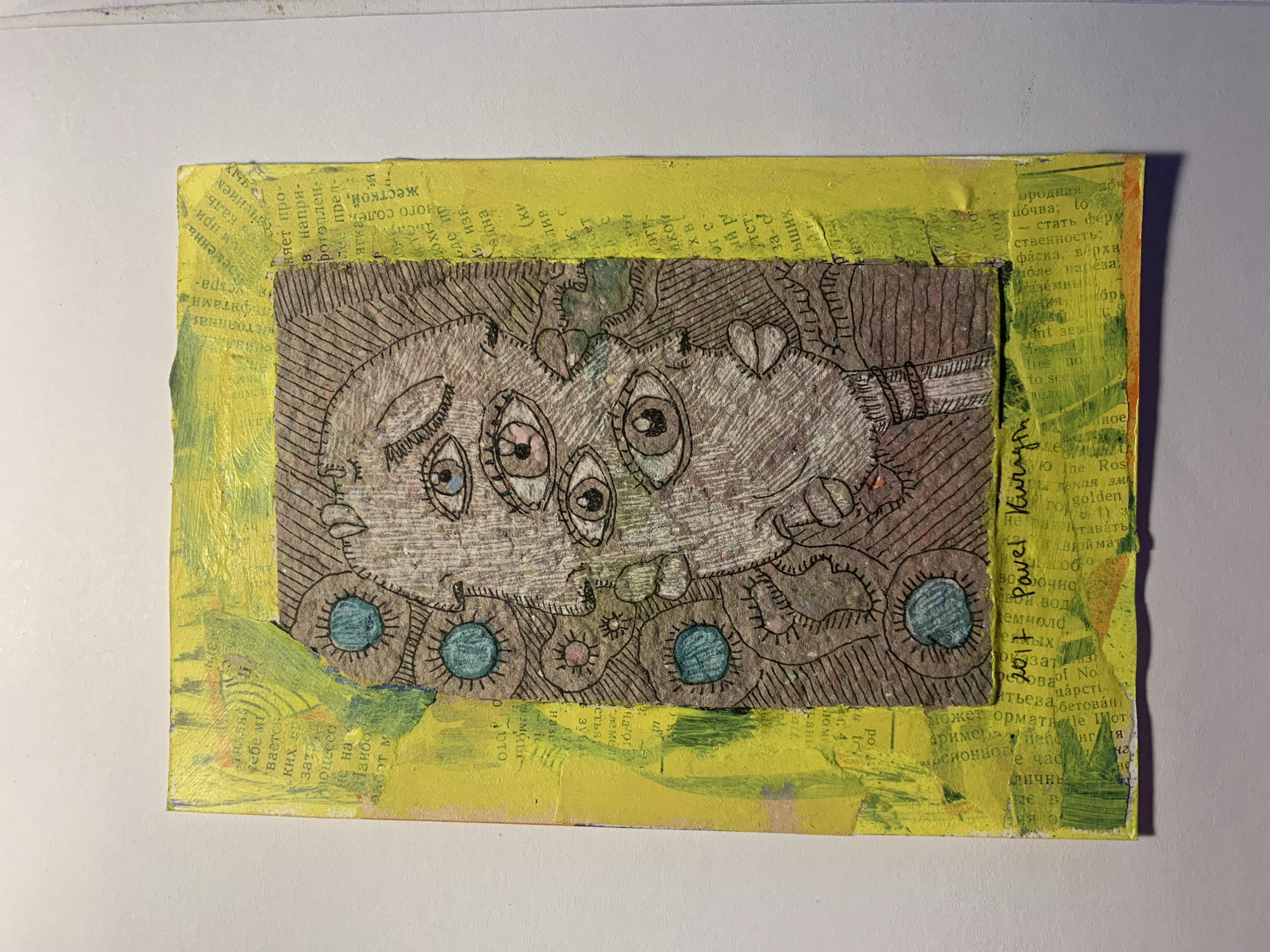 This is a collage artwork on a yellow, textured background. The central focus is a drawing of a creature with multiple eyes, rendered in black ink on brown paper. The creature has a whimsical, slightly unsettling appearance, with a large head and a segmented body. Below the creature, there are several circular shapes, some filled with blue and pink colors. The background is made of torn newspaper fragments, with visible Russian text. There is also some green paint applied in broad strokes around the edges of the collage. The text visible includes phrases like "Golden," "The Ros," and what appears to be Russian Cyrillic. The overall aesthetic is folk art or outsider art, with a handmade and expressive quality. The artwork is signed "Pavel Kirur 2021" on the right side, indicating it was created by an artist named Pavel Kirur in 2021. The location context of Sergeevo, Russia, suggests the newspaper clippings might be from local publications.