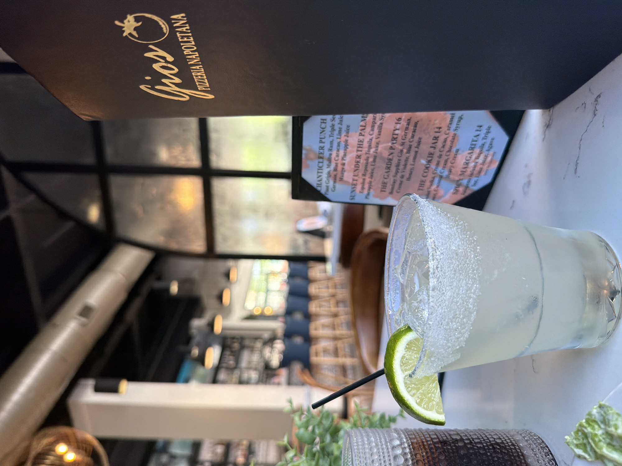 Here's a detailed description of the image:
The image depicts a "Spicy Margarita" on a table, garnished with salt and a lime wedge. The drink sits in front of a menu from "Gios Pizzeria Napoletana" revealing other drink options like "Chanticleer Punch" and "The Cookie Jar." The background shows an indoor-outdoor restaurant setting with visible seating and lighting fixtures. Based on the restaurant name, the photo was likely taken in Myrtle Beach, United States of America. The environment is casual and possibly during the daytime, given the natural light. No people are directly visible in the image, but the presence of the drink and menu suggests it's a customer's view.