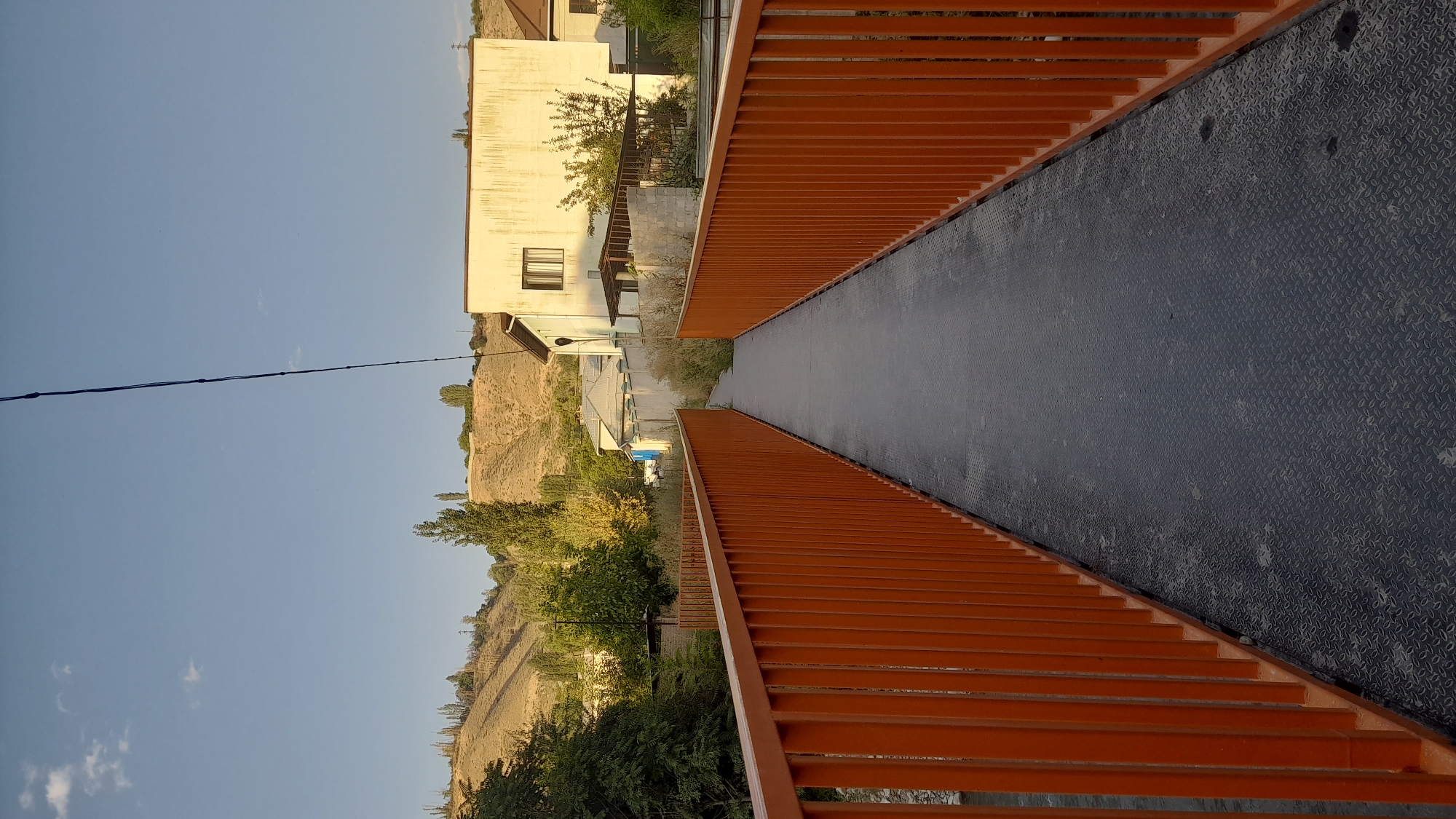 The image shows a pedestrian bridge with orange railings and a dark metal walkway, leading towards a residential area with a large, light-colored building. The bridge appears to be over a body of water, though the water is not clearly visible. The background features a gently sloping, dry hillside dotted with sparse vegetation and trees. A single dark wire extends diagonally across the upper portion of the frame. The sky is clear and blue, with a few small clouds. The scene suggests a rural or semi-rural setting, possibly on the outskirts of a city like Osh City, Kyrgyzstan, given the arid landscape and the style of the buildings. The time of day appears to be late afternoon or early evening, indicated by the warm, low angle of sunlight illuminating the hillside and the building. There are no people or animals visible in the image.