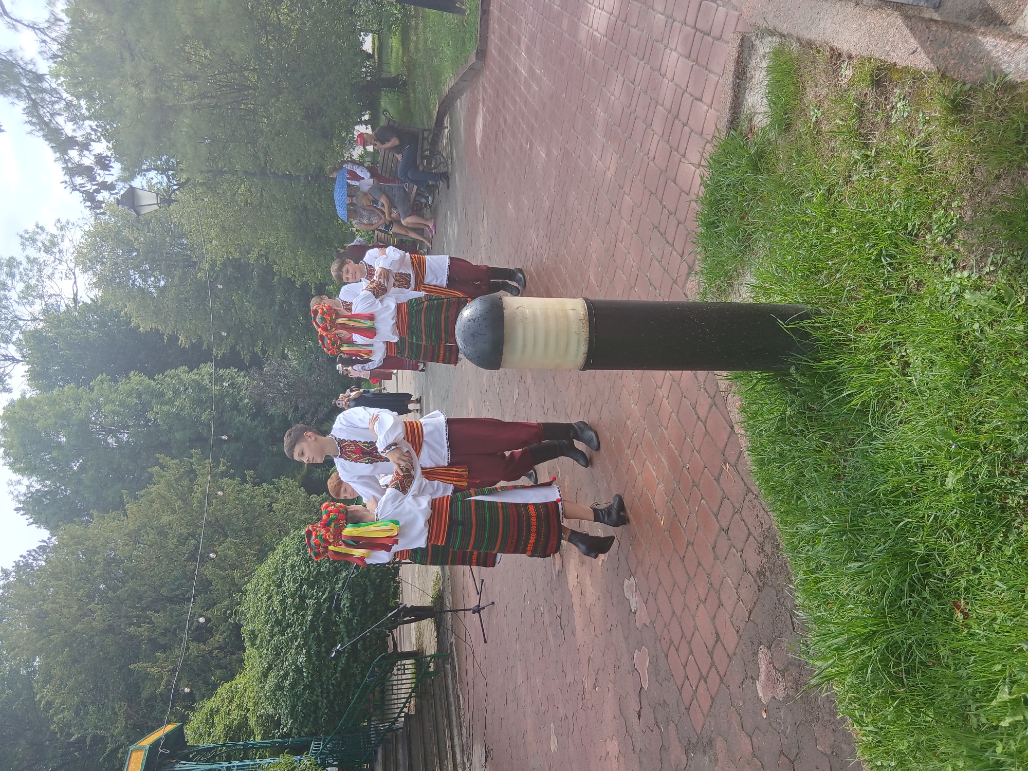 A group of young people in traditional Ukrainian folk costumes are dancing outdoors on a paved walkway. The dancers, consisting of three males and two females, are positioned in the center of the frame. They are adorned in white embroidered shirts, colorful skirts or trousers, and decorative headwear. The female dancers wear elaborate headdresses with ribbons, while the males wear sashes. They appear to be holding hands or interacting as part of a choreographed dance.
In the background, an audience is seated on benches under a canopy of trees. Some individuals are holding umbrellas, suggesting a sunny but potentially warm day. A lamppost is visible in the mid-ground, and a microphone stand is set up near the dancers. The walkway is paved with red bricks, and grass borders the scene. The environment suggests a public park or an outdoor event space. The scene takes place in Truskavets, Ukraine, a city known for its spa resorts and natural beauty.